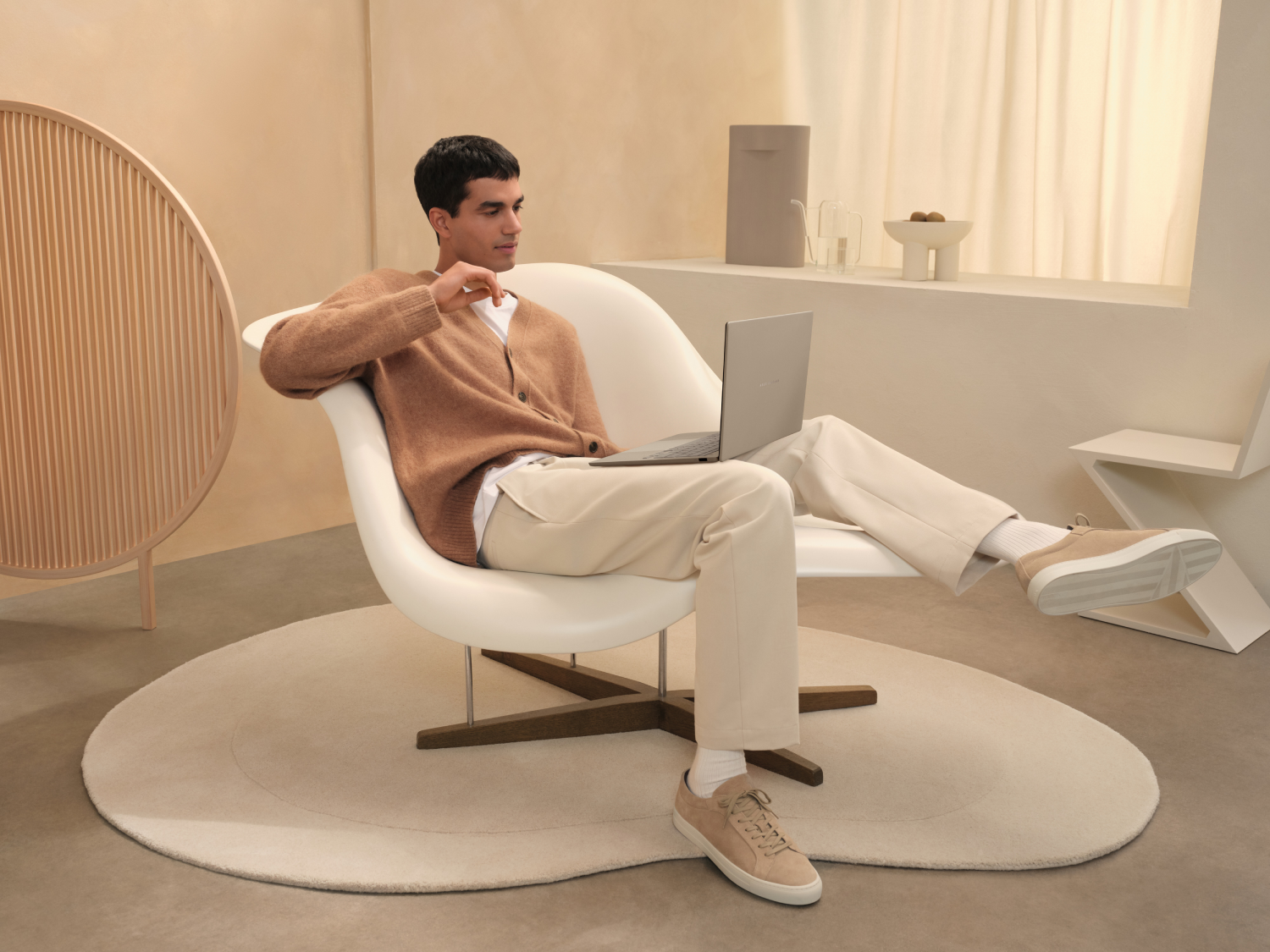 Un hombre está sentado en una silla blanca usando una laptop. Lleva un suéter marrón y pantalones beige. La habitación tiene colores claros.