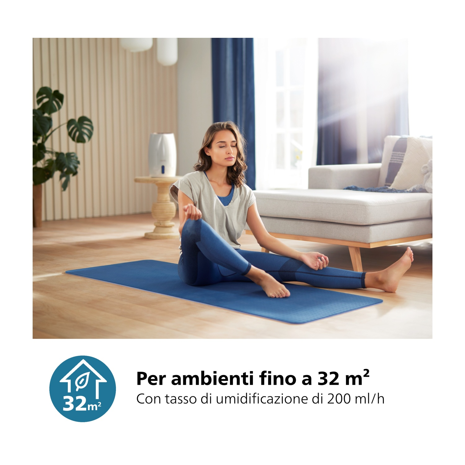Una donna seduta su un tappetino blu, con gli occhi chiusi. È in una stanza con un divano, una finestra e un umidificatore.