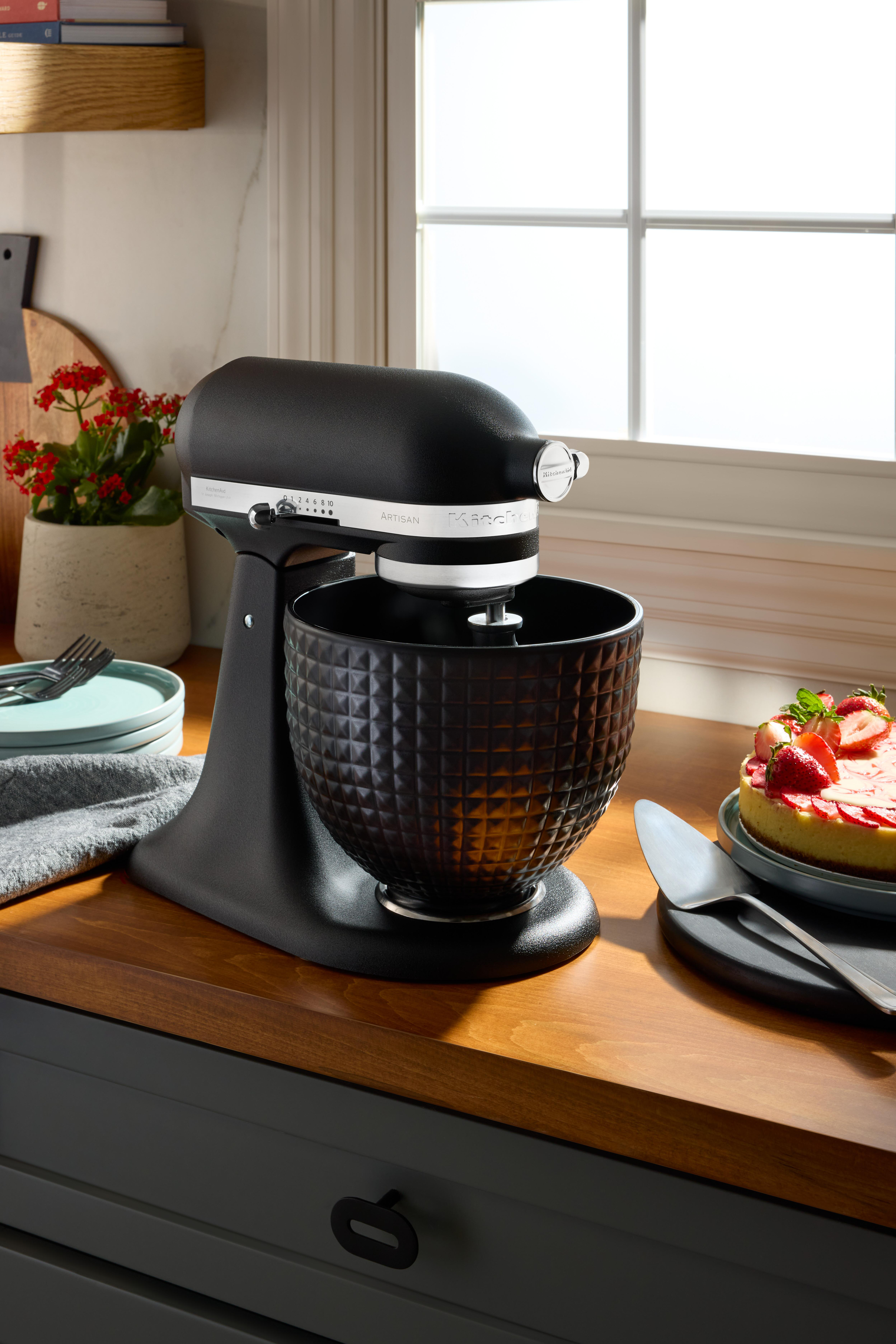 Schwarzer KitchenAid-Mixer mit strukturierter Schüssel auf einer Holztheke neben einem Fenster und einem Kuchen.