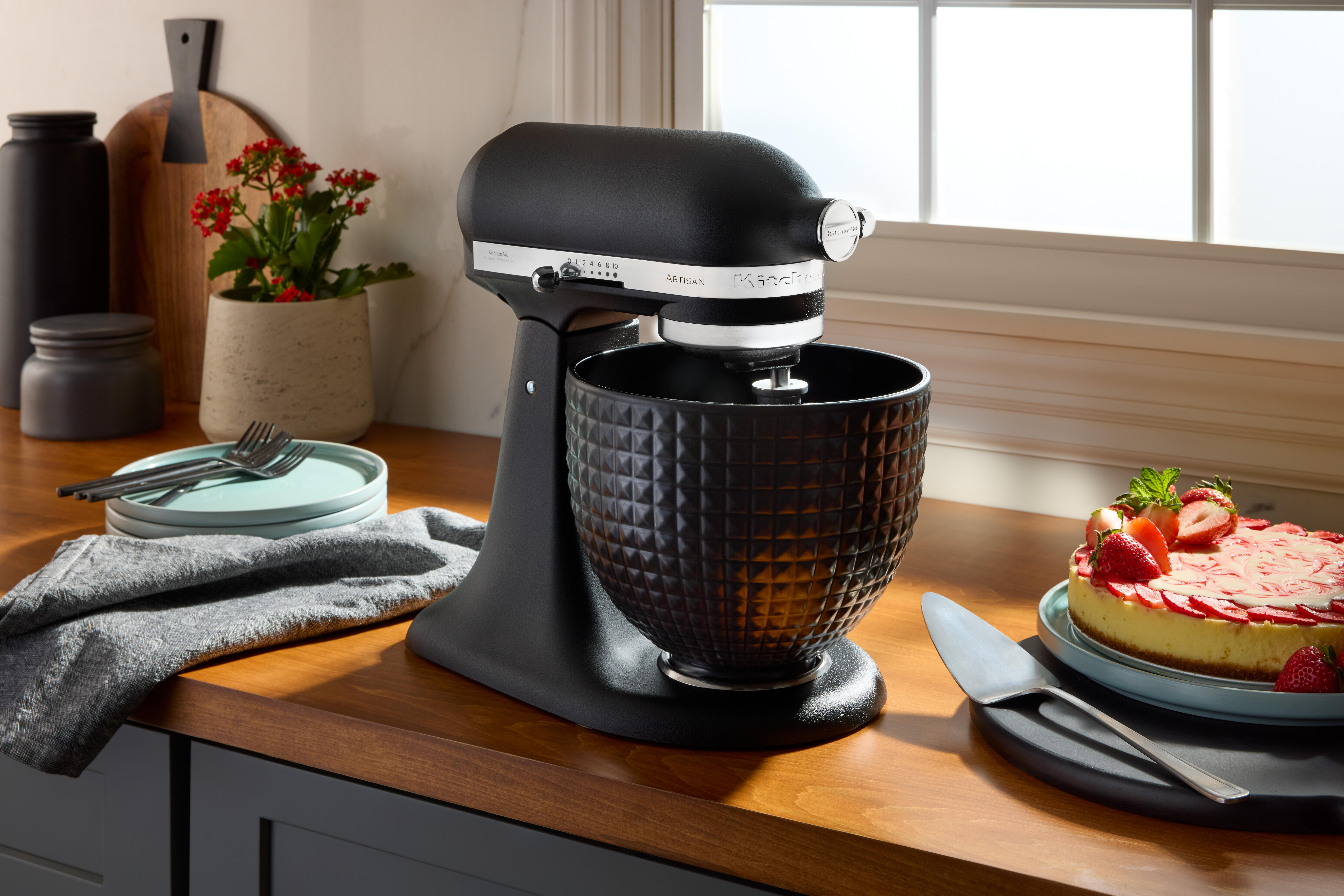 Schwarzer KitchenAid-Mixer, Kuchen, Teller, Gabeln und Pflanzen auf einer Holztheke, beleuchtet von Tageslicht.