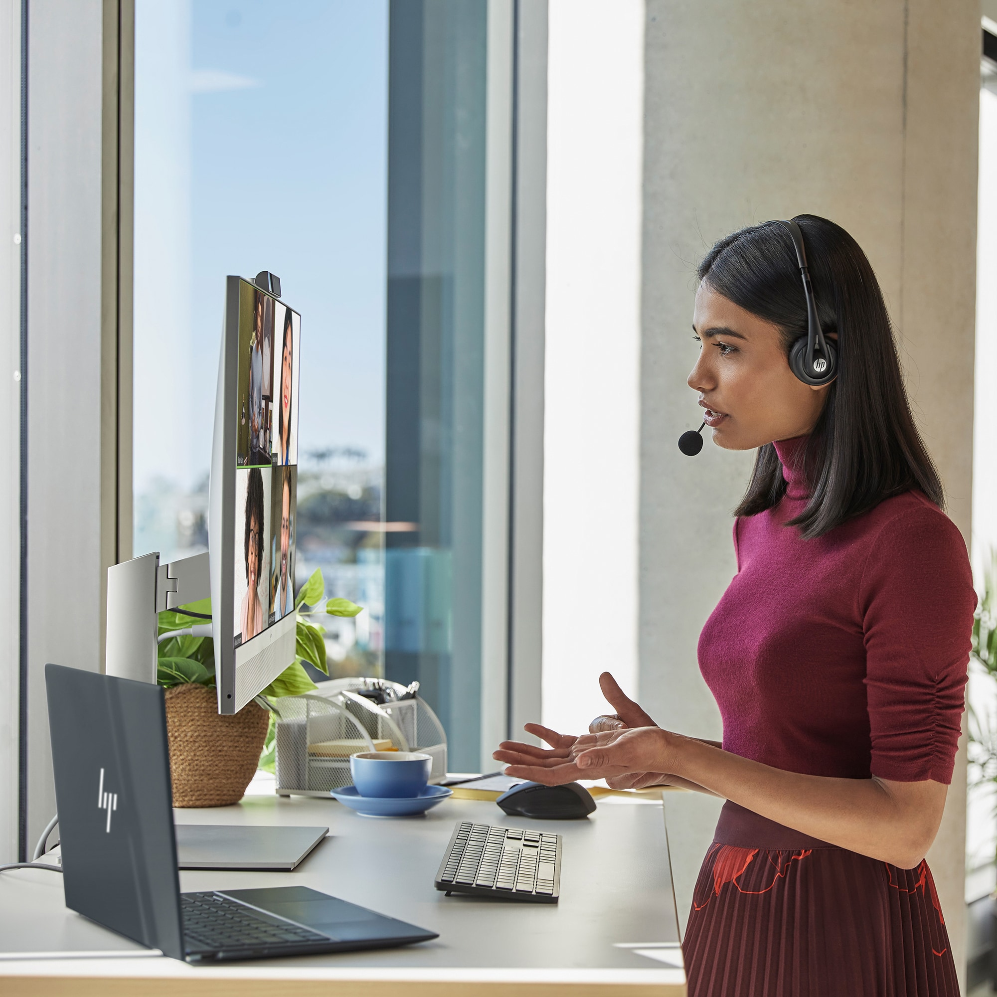 Frau mit Headset während eines Videoanrufs. Monitor zeigt andere Teilnehmer. Laptop und Tastatur auf dem Schreibtisch. Fenster im Hintergrund.