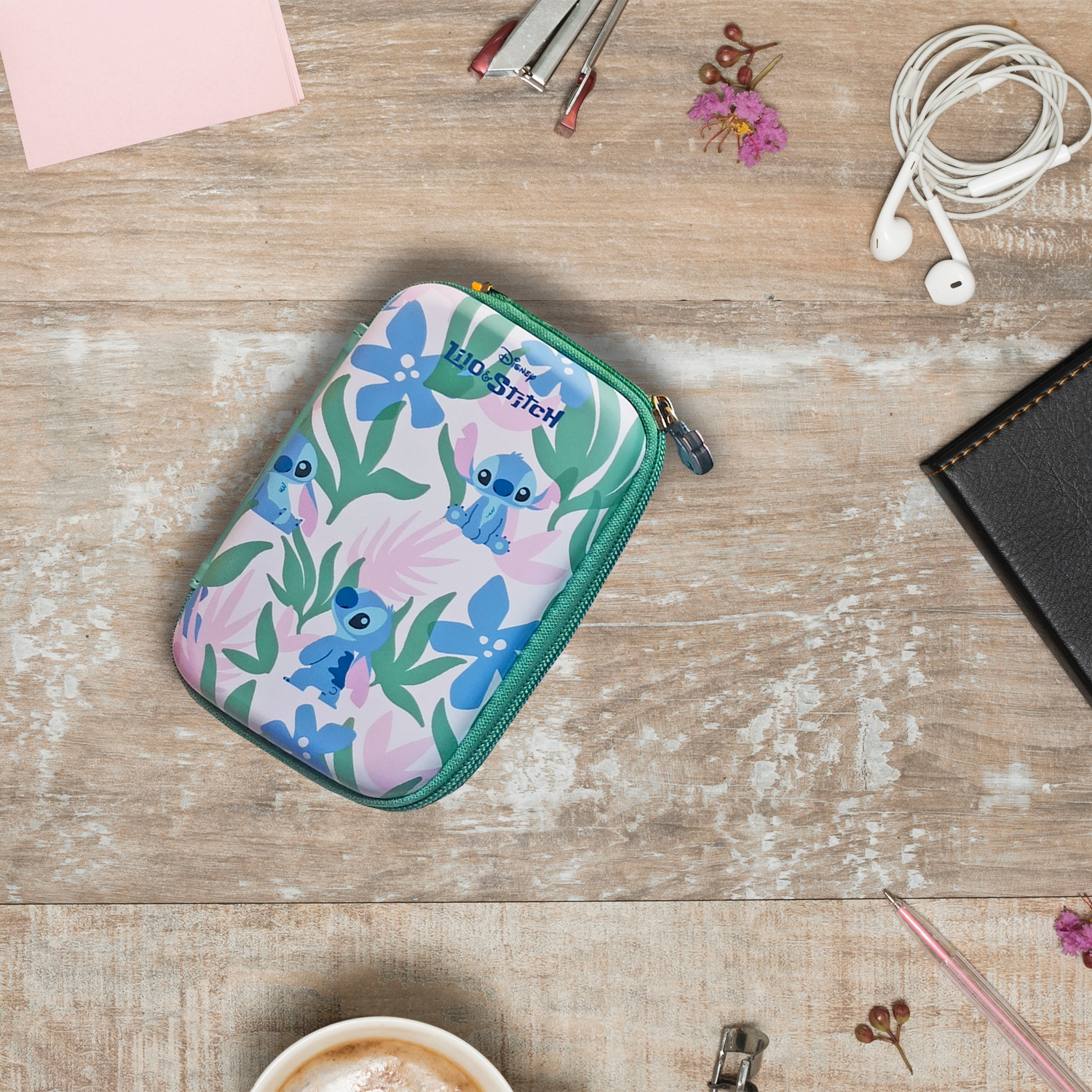 Un estuche de Disney Stitch sobre una mesa de madera con flores, grapadora, auriculares, bolígrafo y cuaderno.