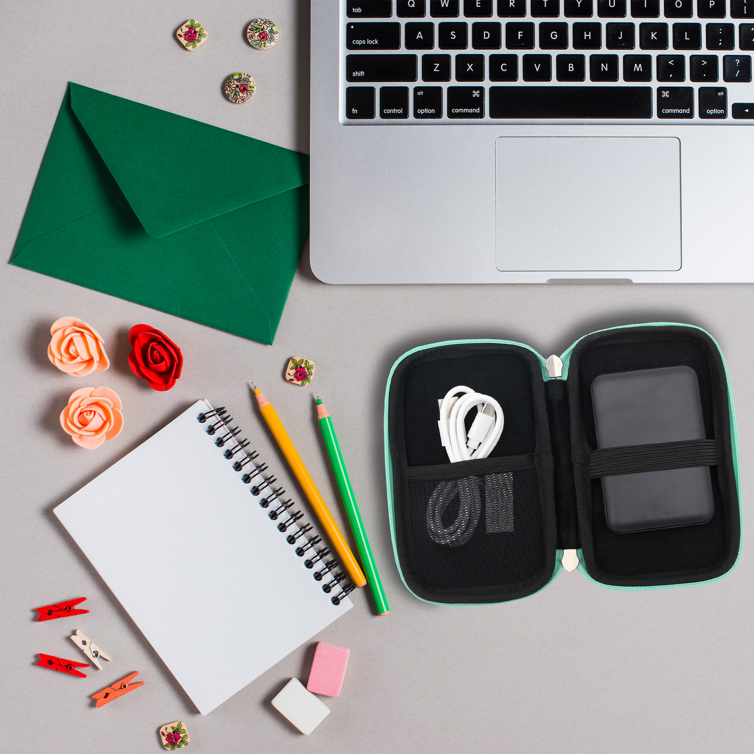 Ordenador portátil, estuche abierto con disco duro y cables, sobre verde, bloc de notas, lápices, gomas de borrar y elementos decorativos sobre superficie gris.