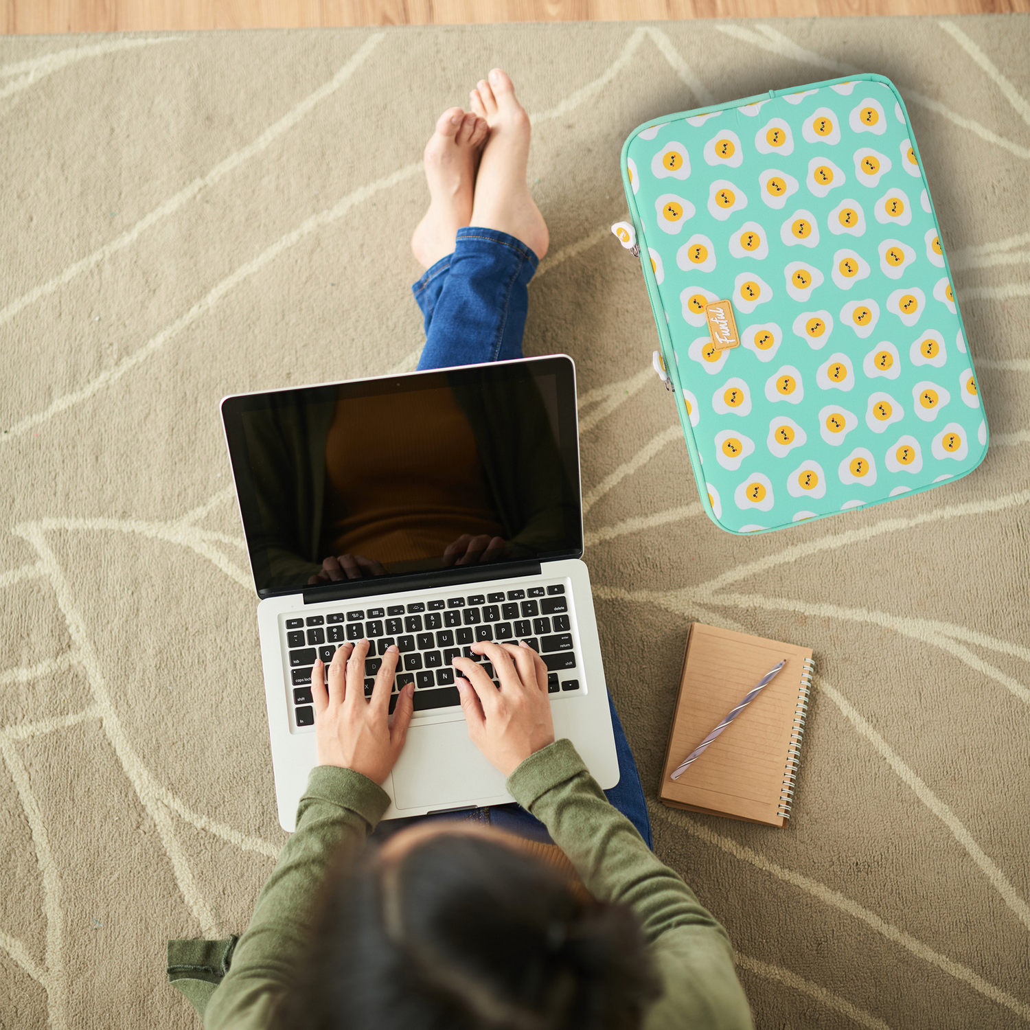 Persona sentada en una alfombra, usando una laptop. Una funda con estampado de huevos fritos está al lado. También se ven un cuaderno y un bolígrafo.