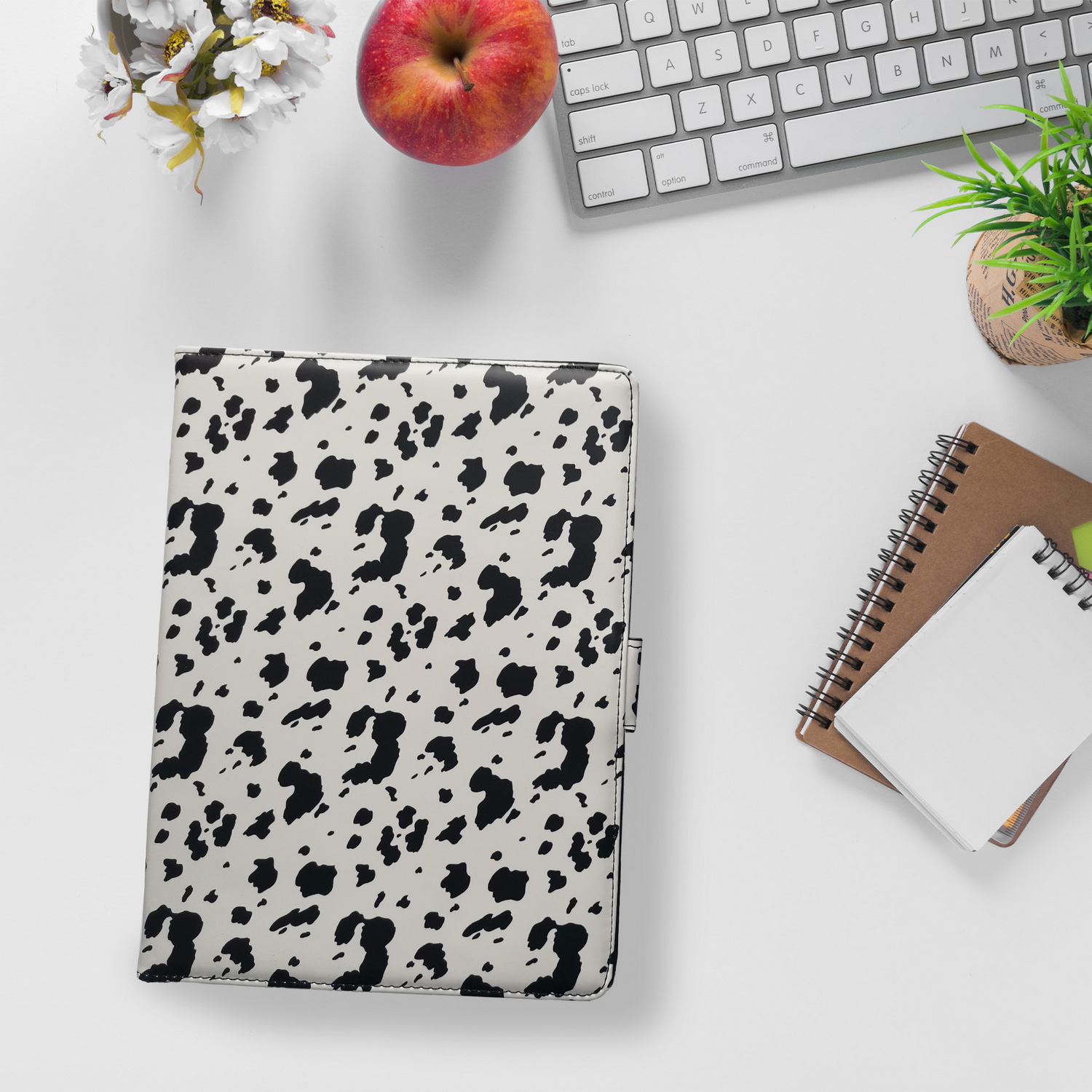 Un escritorio blanco con una carpeta manchada en blanco y negro, una manzana roja, un teclado, una planta pequeña y cuadernos.