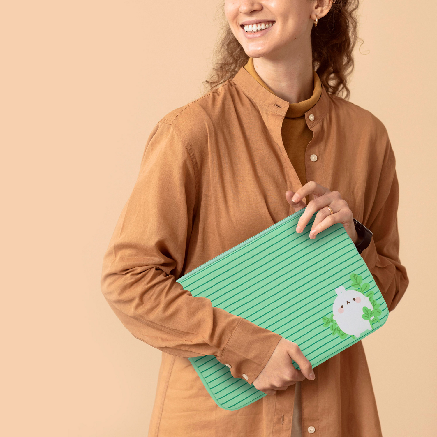 Una mujer sostiene un cuaderno verde con una ilustración de conejito. Viste una camisa marrón y sonríe. El fondo es color melocotón neutro.