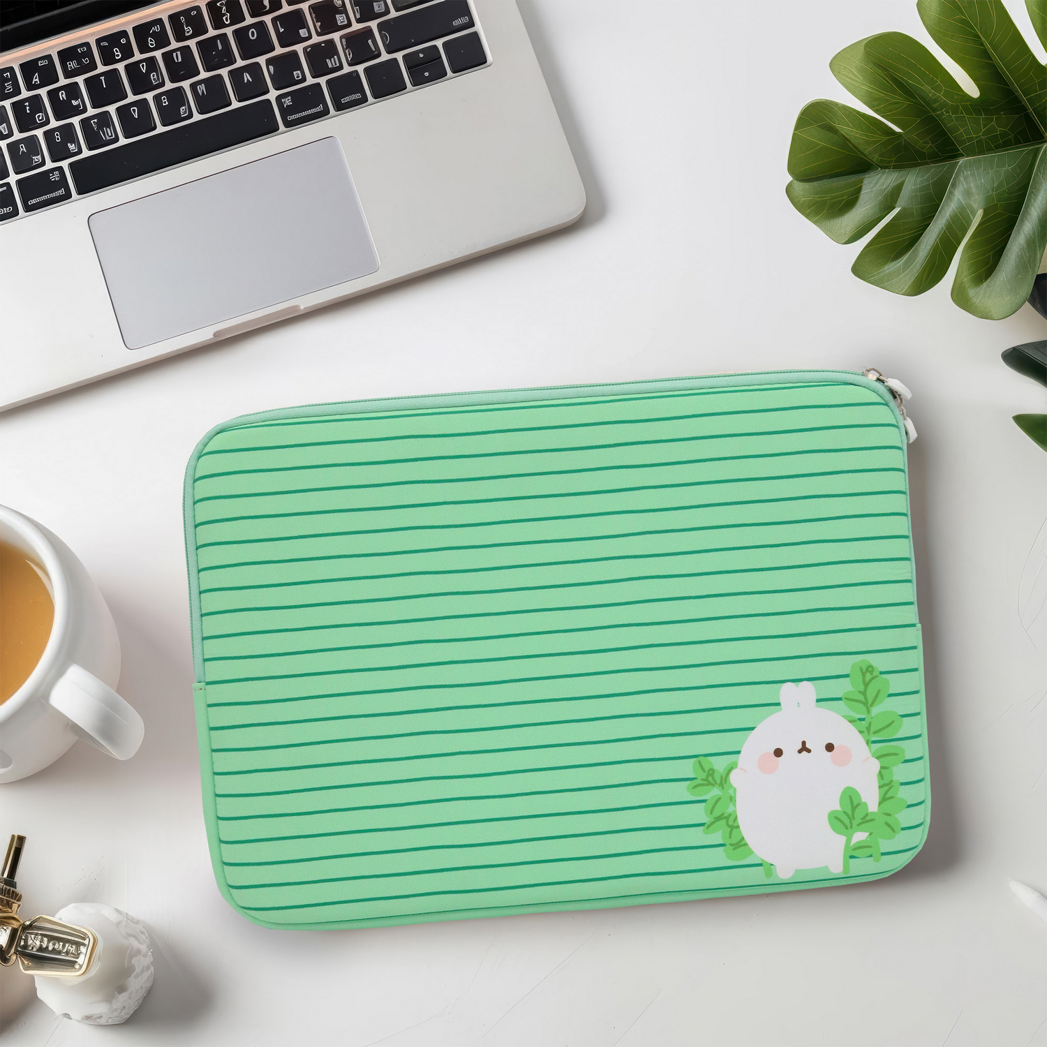 Laptop, funda a rayas verdes con conejito de dibujos animados, taza de café y hojas verdes sobre superficie blanca.