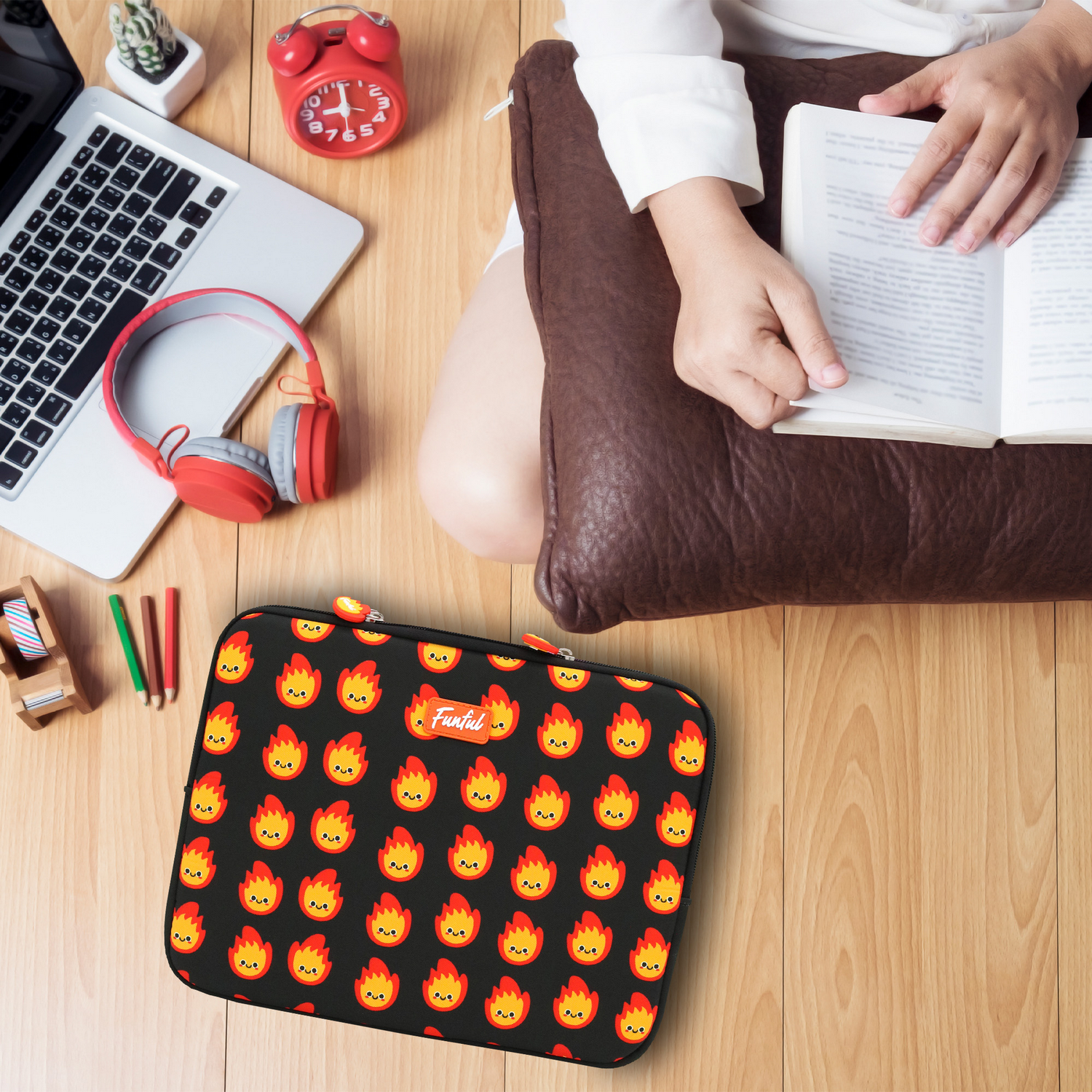 Una persona leyendo un libro, rodeada de un portátil, auriculares, un reloj y una funda con un diseño de llamas, todo sobre madera.