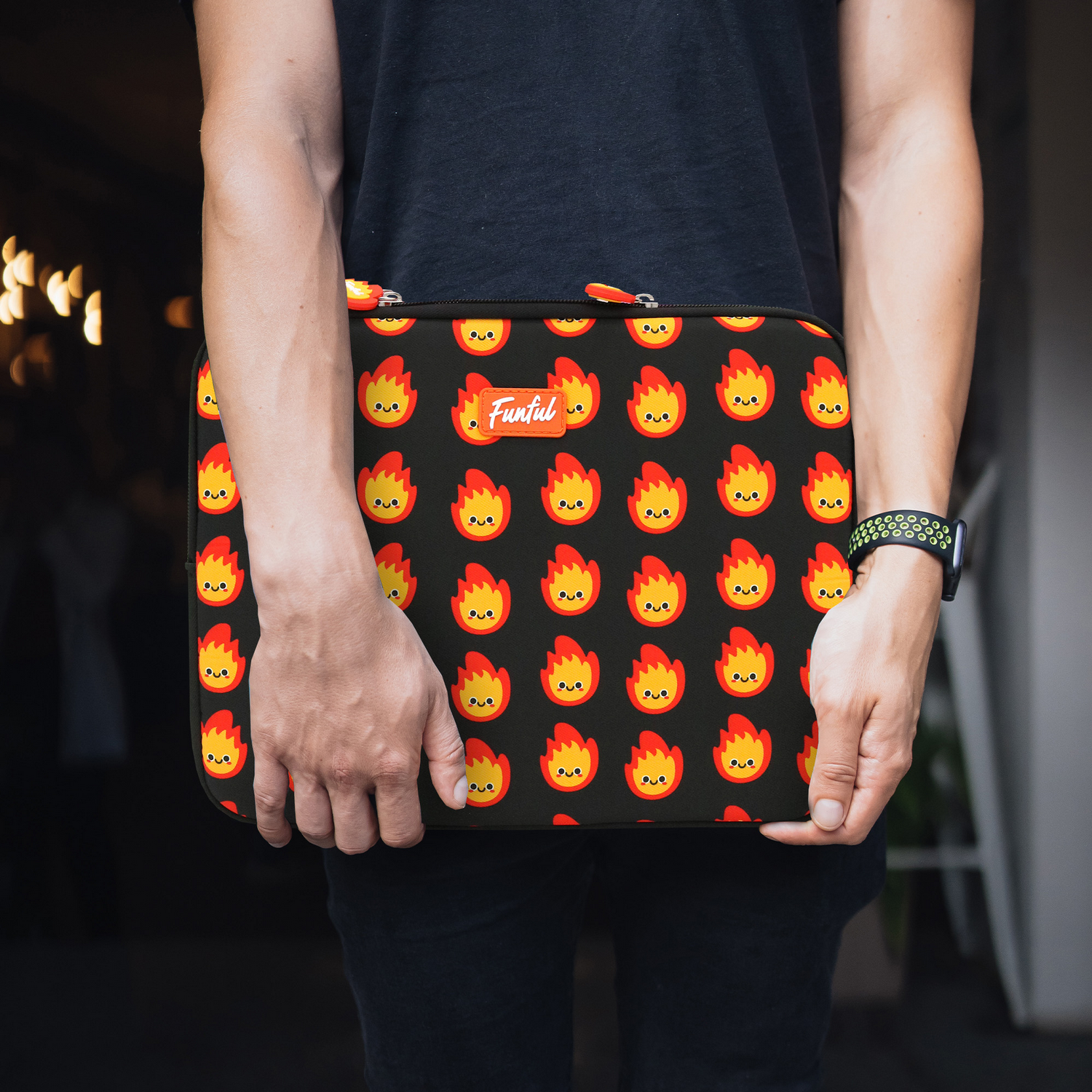 Una persona sostiene una funda para portátil con un fondo negro y gráficos de fuego de dibujos animados repetidos. La persona lleva una camisa negra.