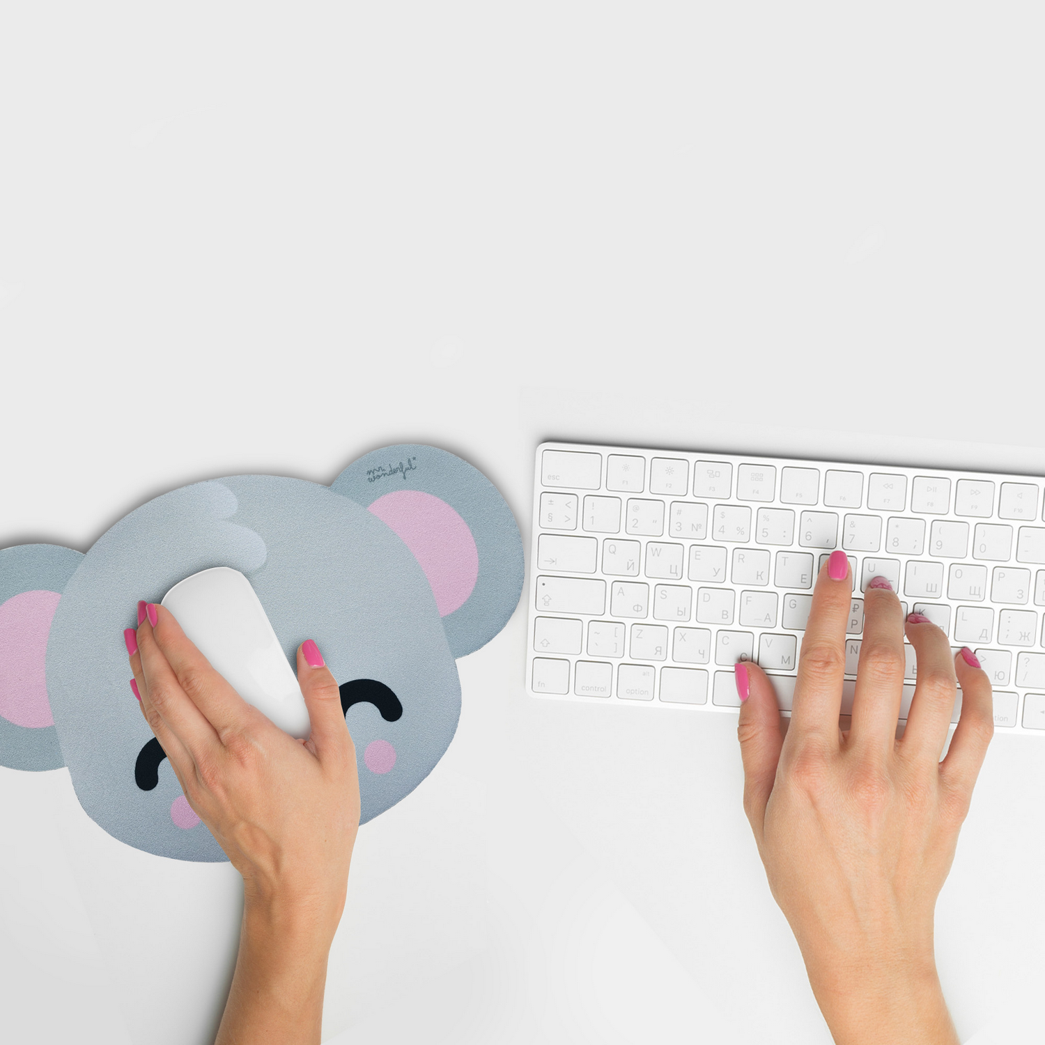 Un escritorio con un mousepad de koala, teclado blanco y manos escribiendo. Uñas rosas en ambas manos.