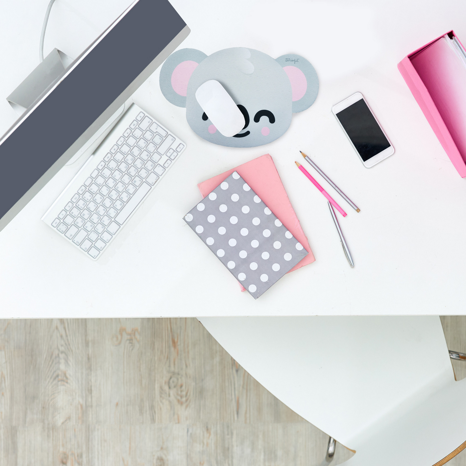 Escritorio blanco con computadora, teclado, alfombrilla de ratón koala, teléfono, cuadernos, lápices y una caja de almacenamiento rosa. Suelo de madera y silla visibles.