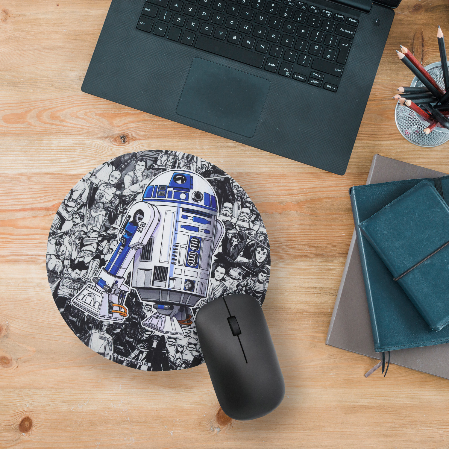 Un escritorio con una laptop, un ratón, un mousepad de Star Wars con R2-D2, lápices y dos cuadernos sobre una superficie de madera.