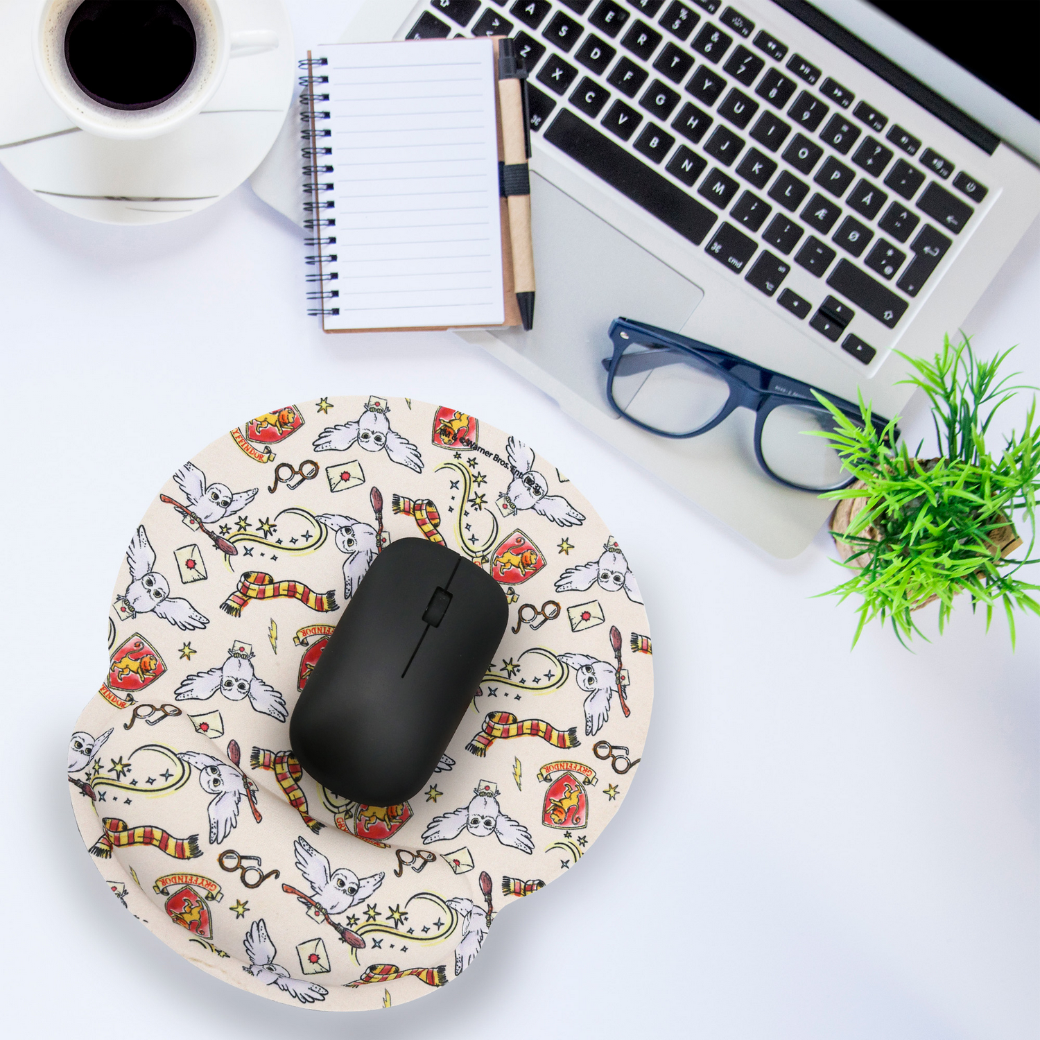 Un escritorio con una laptop, gafas, un bloc de notas, una taza de café y un ratón sobre una alfombrilla de ratón de Harry Potter.