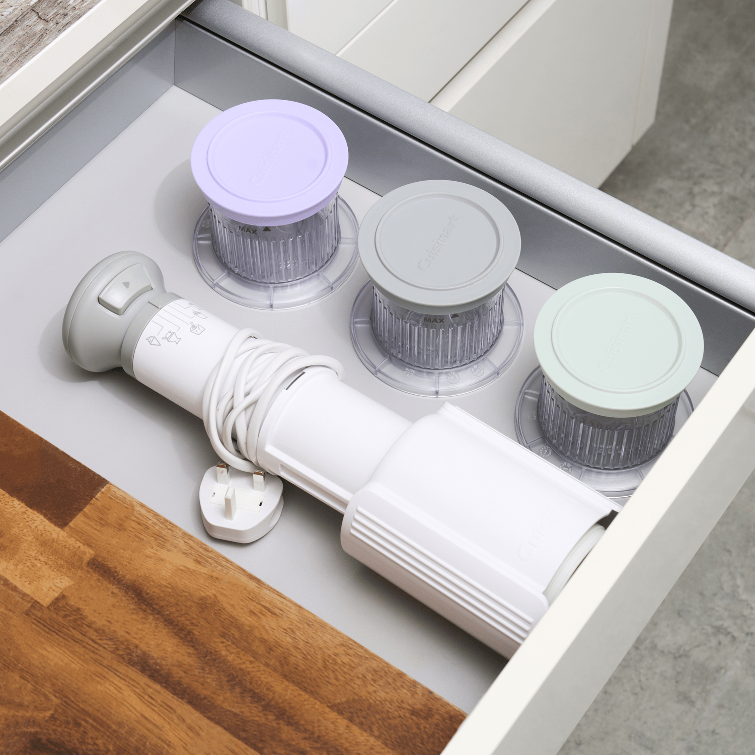 Een lade bevat een witte staafmixer, drie voedselverwerkerskommen met deksels en een houten aanrecht.