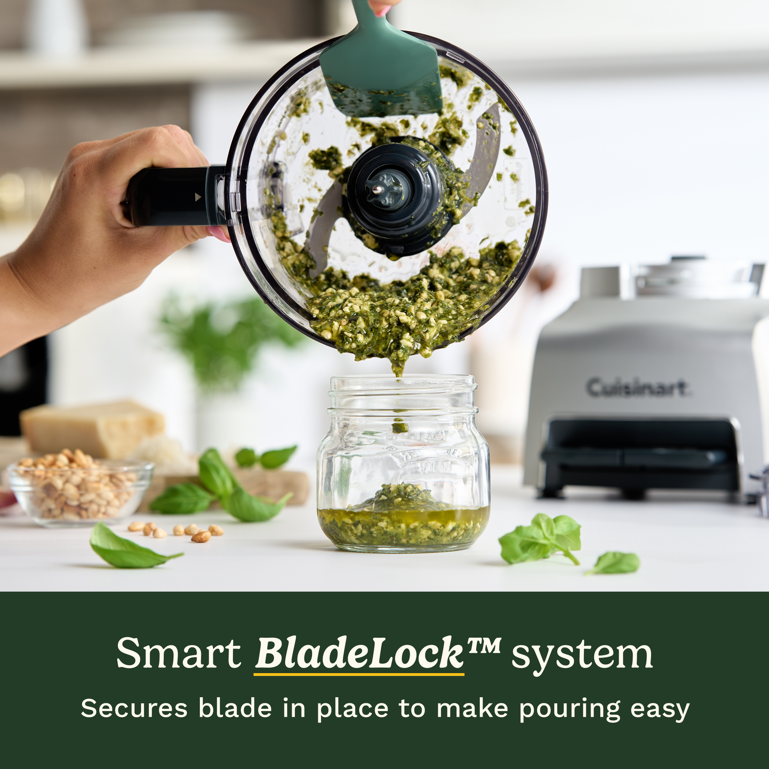 A food processor pours pesto into a jar. Basil leaves, nuts, and ingredients are on the table. A Cuisinart is in the background.