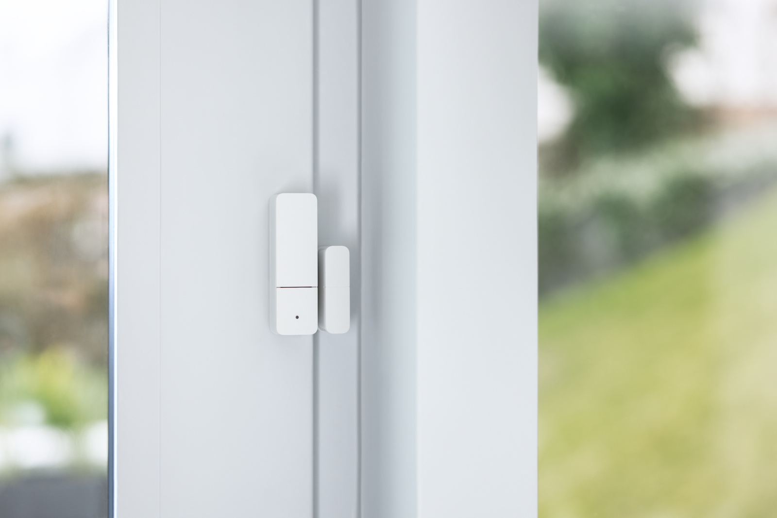 Ein weißer Fenstersensor ist an einem weißen Türrahmen montiert. Durch das Fenster ist ein verschwommener grüner Rasen zu sehen.