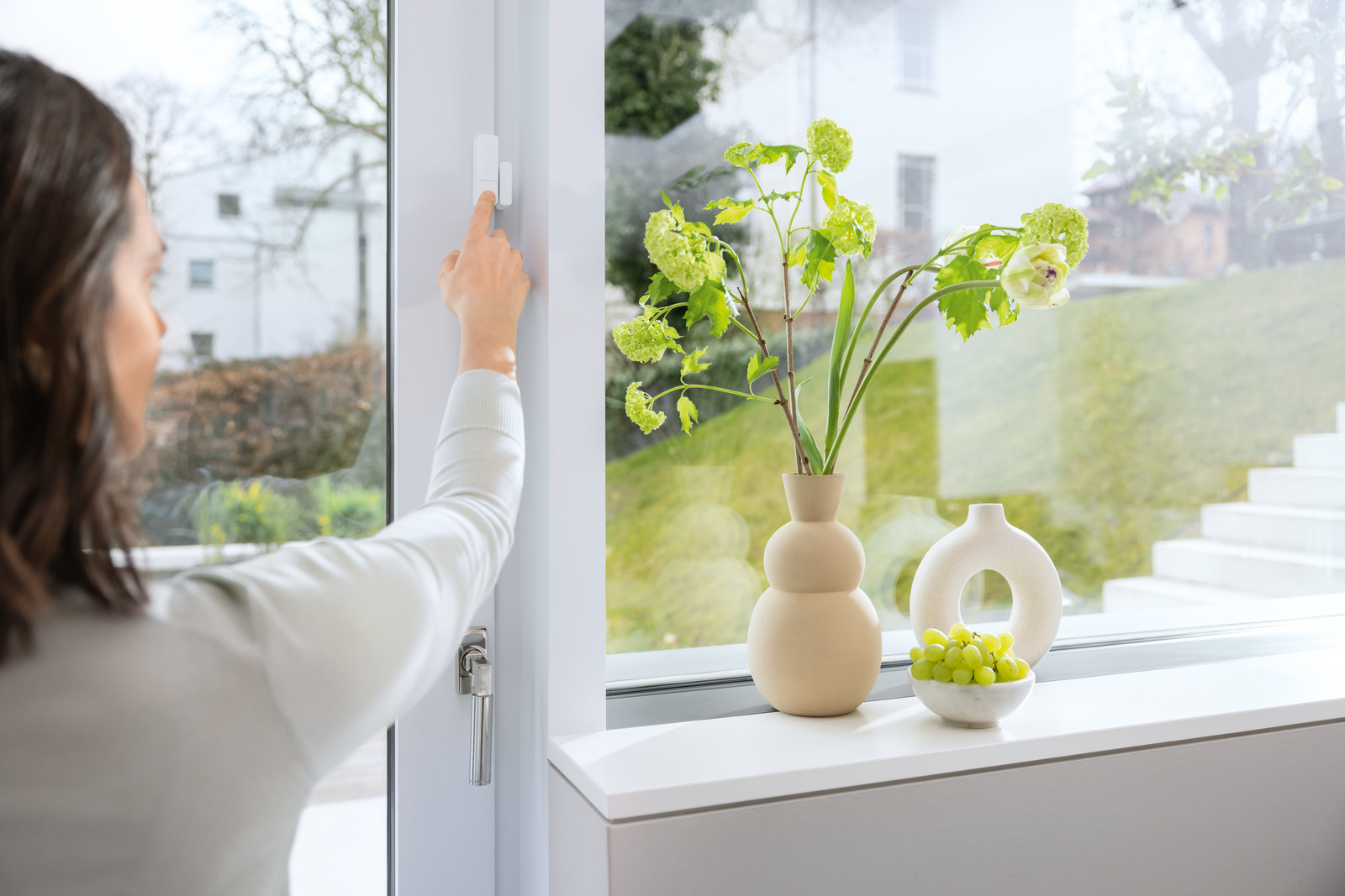 Die Hand einer Frau drückt einen weißen Sensor am Fensterrahmen. Eine Vase mit Blumen und Trauben steht auf der Fensterbank, ein Garten ist sichtbar.