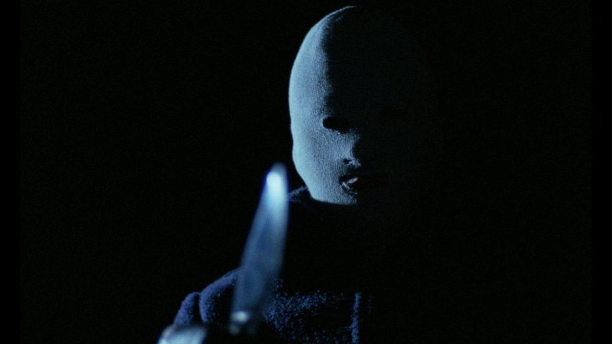 Eine Person mit Maske hält ein Messer in einer dunklen Umgebung. Nur das Gesicht und das Messer sind sichtbar. Der Hintergrund ist schwarz.