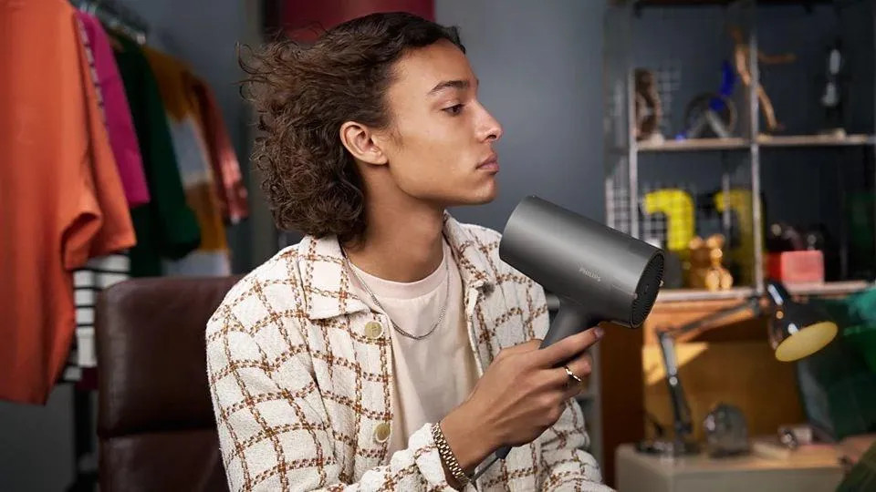 Młody mężczyzna z kręconymi włosami używa szarej suszarki do włosów. Ma na sobie kratkowaną kurtkę i łańcuszek. Tło obejmuje ubrania i półkę.