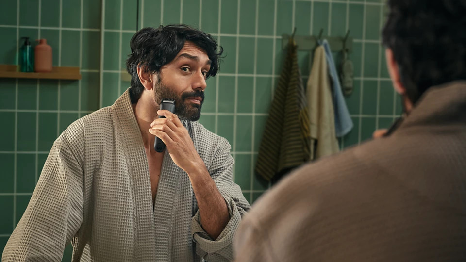 Mężczyzna w szlafroku goli brodę elektryczną maszynką w łazience. Zielone kafelki w tle.