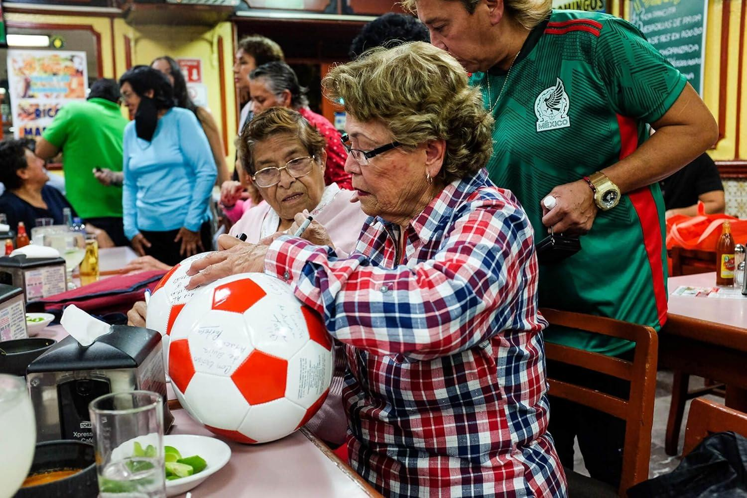 Leute unterschreiben einen Fußball. Eine Frau in einem karierten Hemd und einer Brille unterschreibt. Andere schauen in einem Restaurant zu.