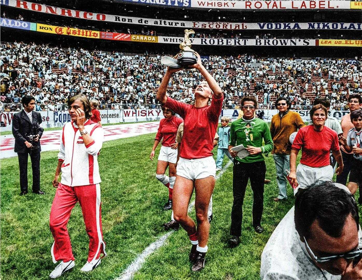 Ein Fußballspieler in roter Hose und Trikot hält eine Trophäe auf einem grünen Feld hoch. Andere Leute umgeben ihn, und Fans füllen die Tribünen.