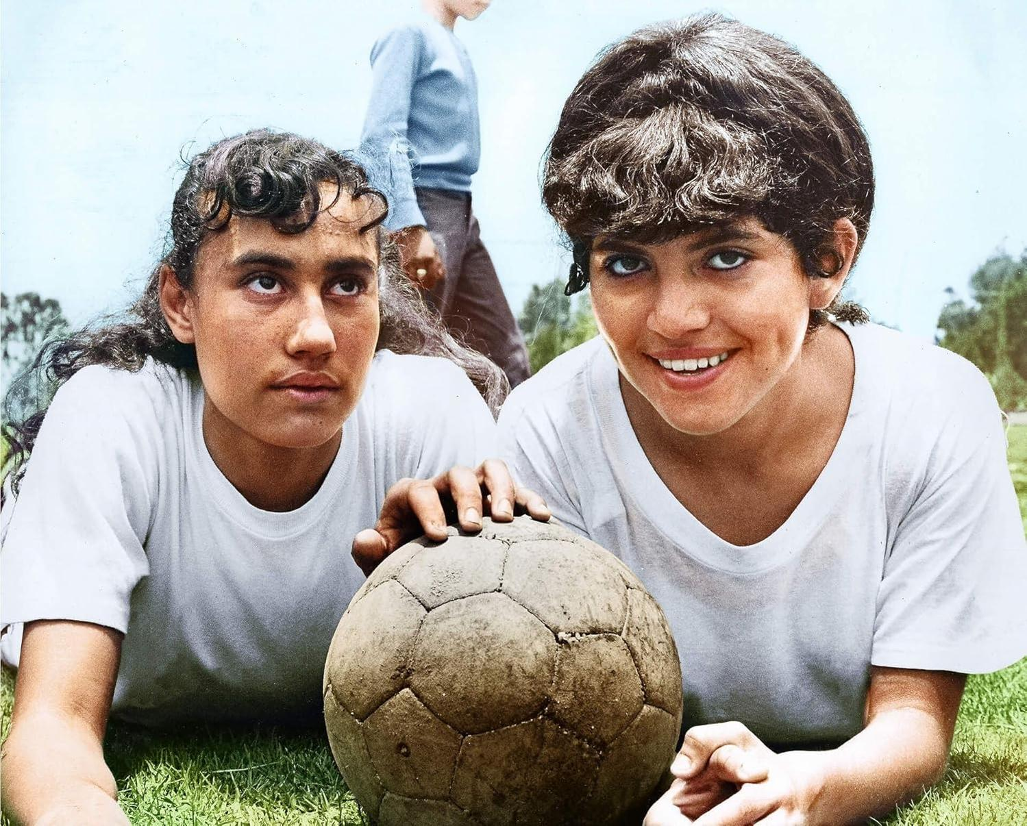Zwei Mädchen auf Gras mit einem Fußball, lächelnd. Ein Junge steht im Hintergrund. Alle tragen weiße Hemden.
