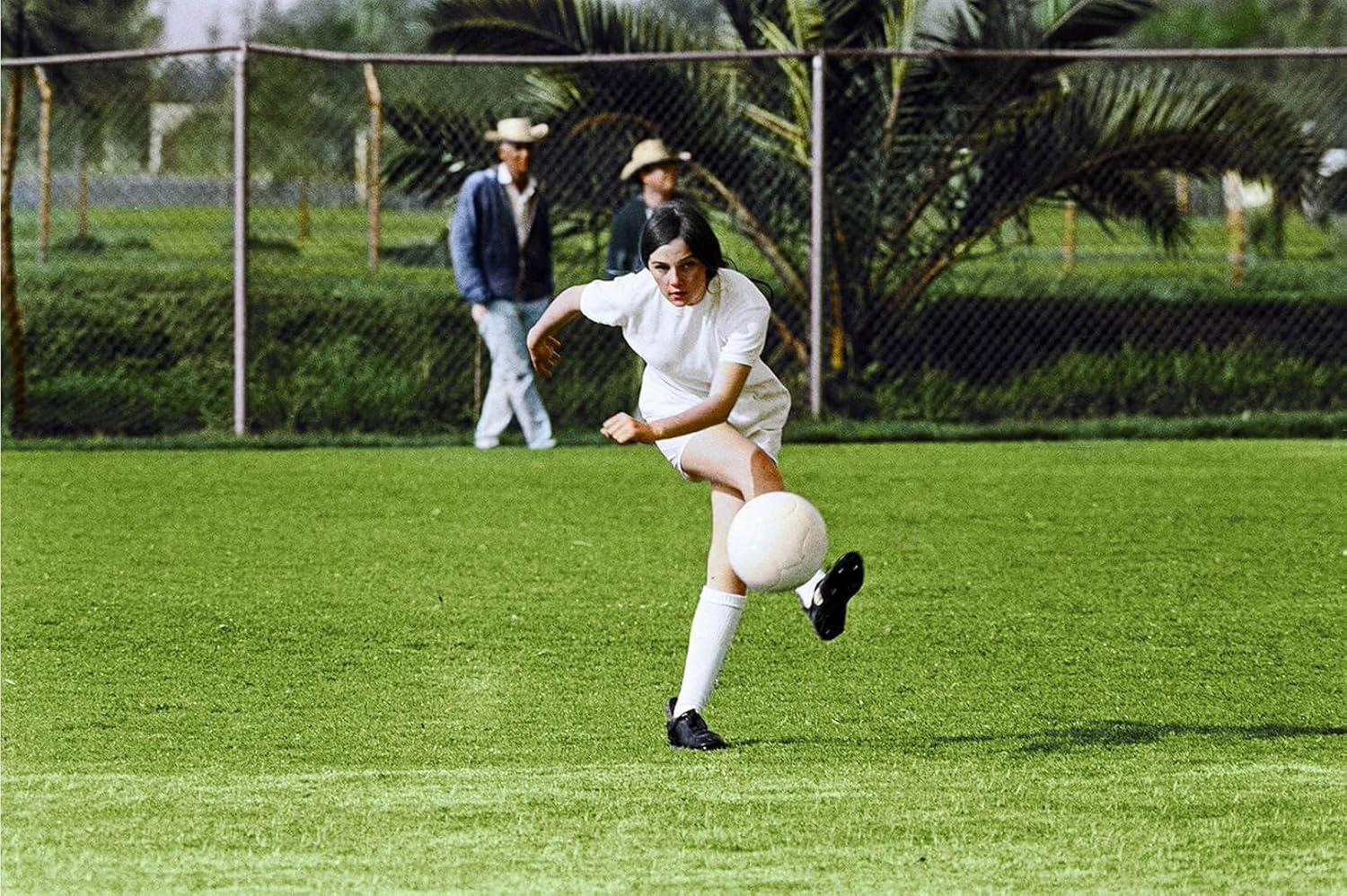 Eine Frau in Weiß tritt einen Fußball auf einem grünen Feld. Zwei Männer stehen im Hintergrund hinter einem Zaun.