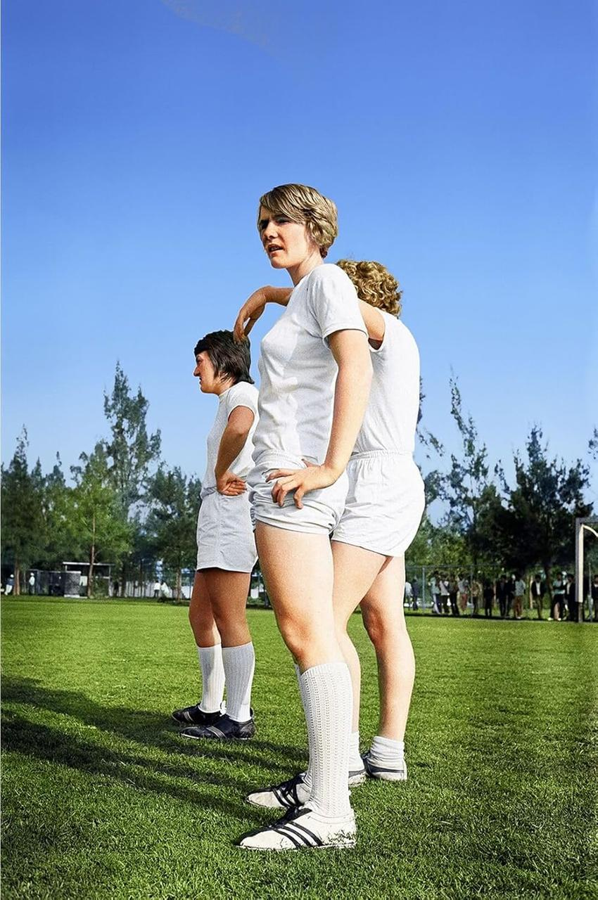 Drei Frauen in weißen Sportoutfits stehen auf einem grünen Feld unter blauem Himmel. Bäume und Menschen sind im Hintergrund.