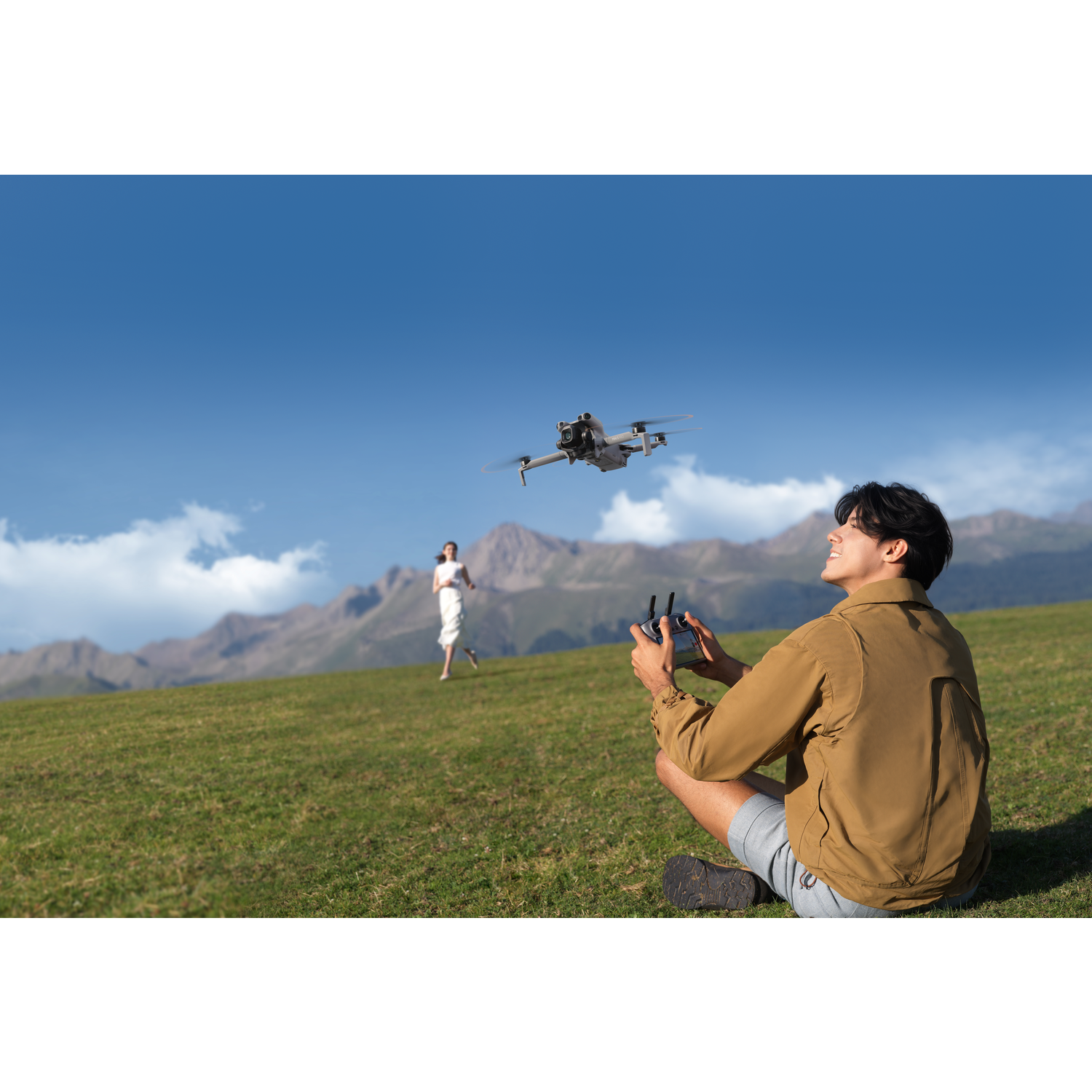 A man operates a drone on a grassy hill. A woman runs in the background. Mountains and a blue sky are in the background.