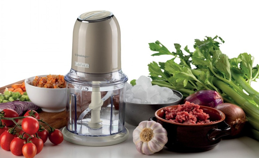 Elektryczny rozdrabniacz do żywności z warzywami, mięsem, lodem i czosnkiem na białej powierzchni. Posiekane warzywa są w misce.