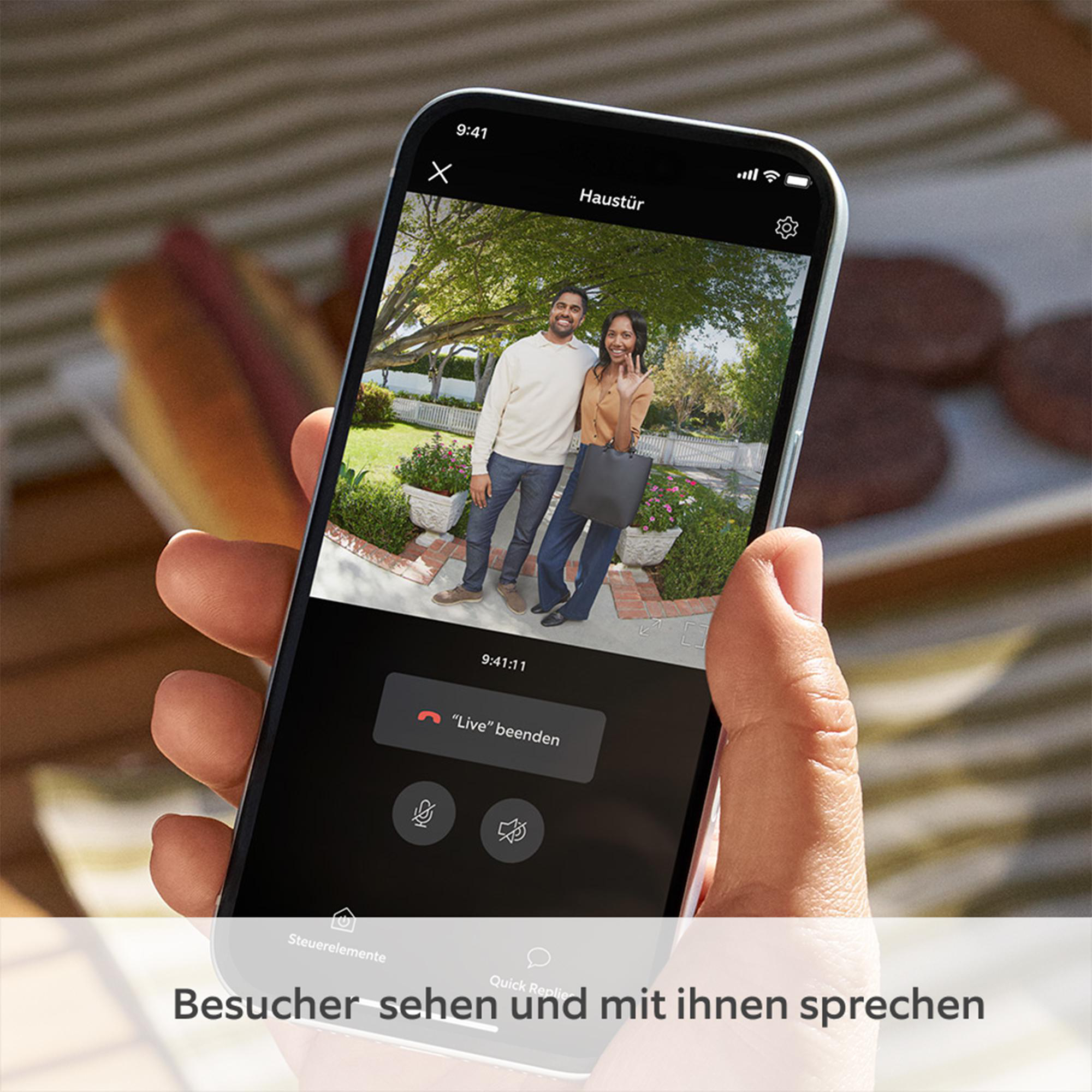 Eine Person hält ein Telefon, das eine Live-Ansicht eines Paares zeigt. Das Telefon zeigt den Text 'Haustür' und 'Live beenden'.