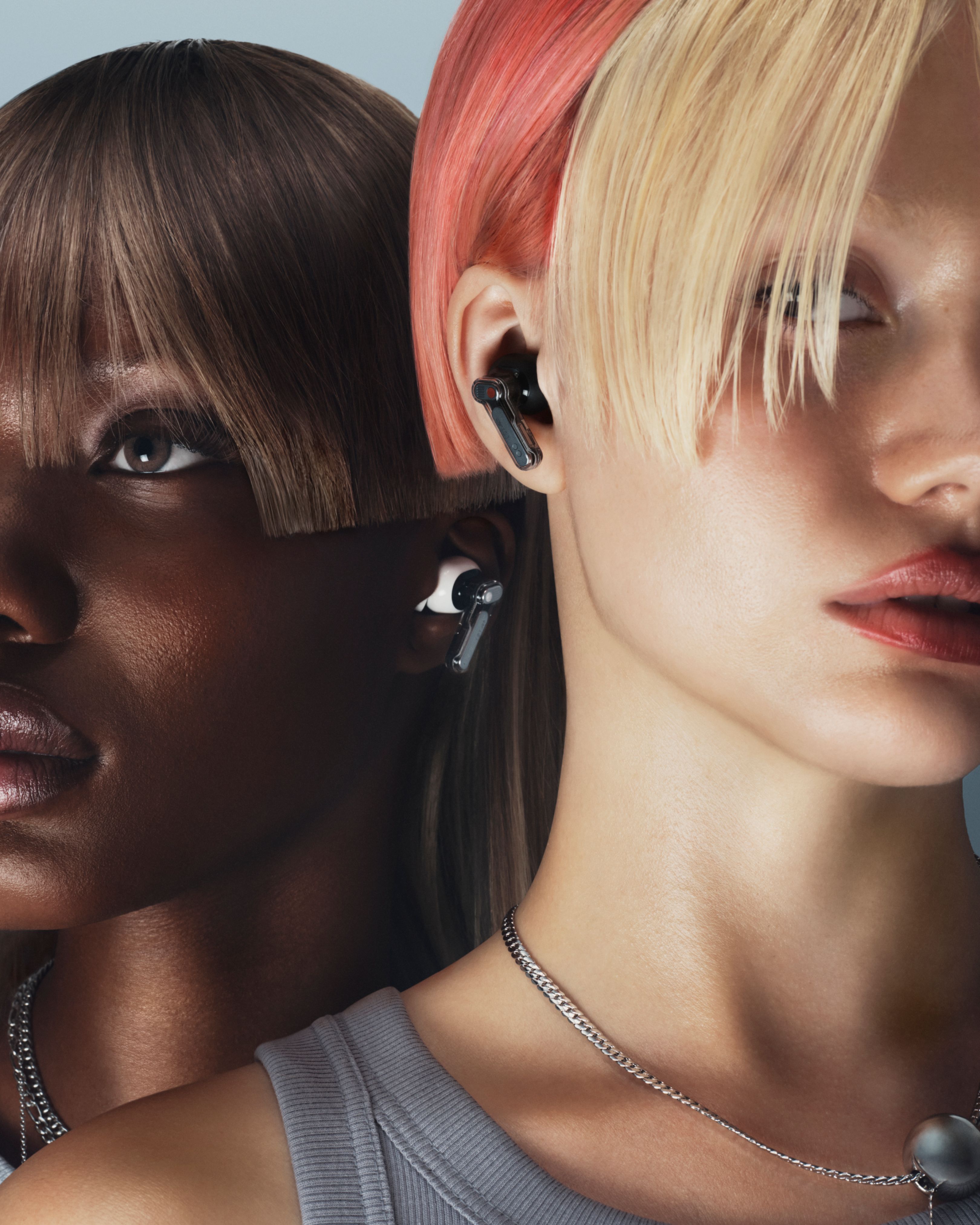 Zwei Frauen mit unterschiedlichen Hauttönen und Frisuren. Eine hat rosa und blonde Haare, die andere braune Haare. Beide tragen Ohrhörer und Halsketten.