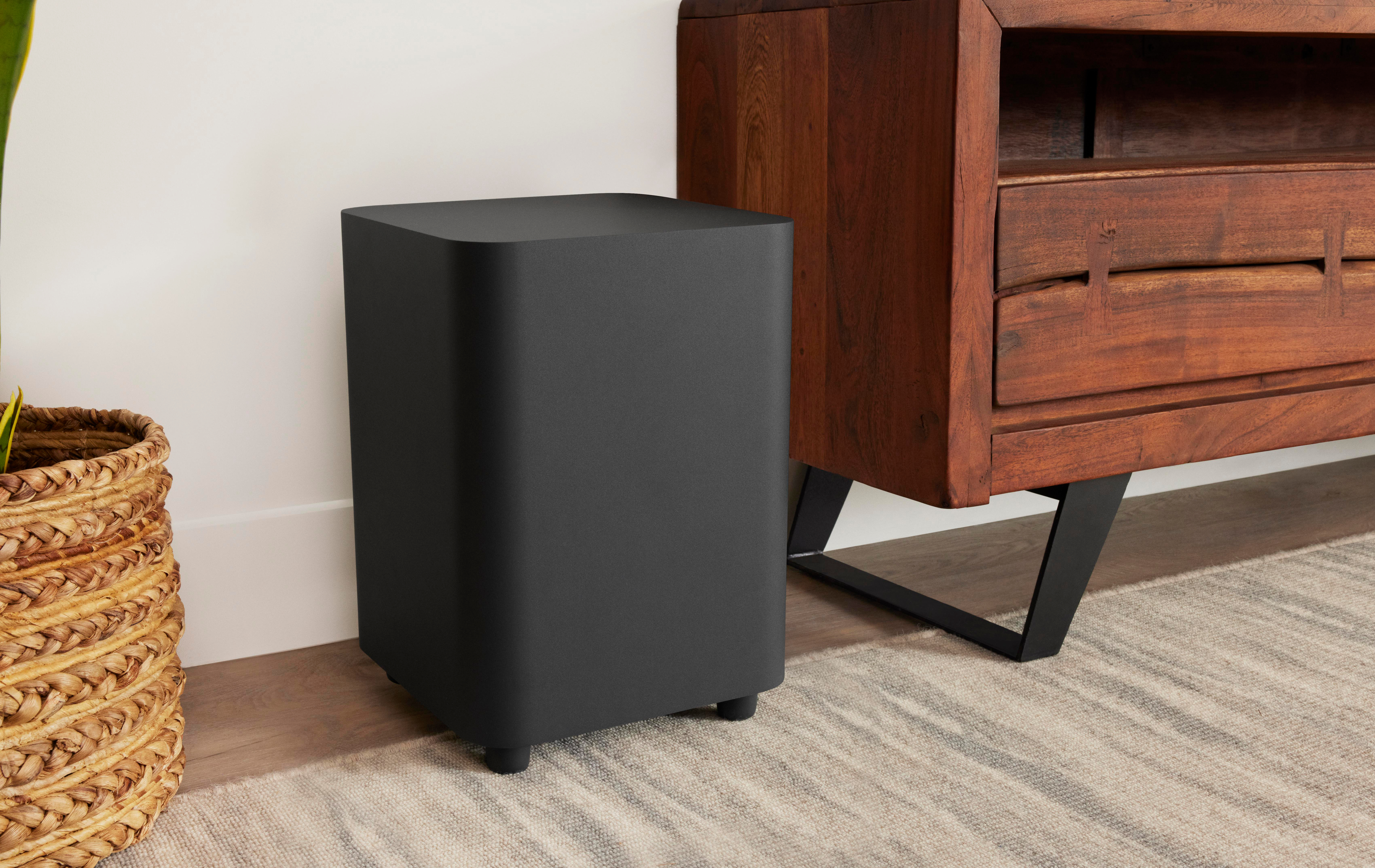 Ein schwarzer Subwoofer steht neben einem Holzschrank mit schwarzen Beinen. Links befindet sich ein geflochtener Korb mit einer Pflanze.