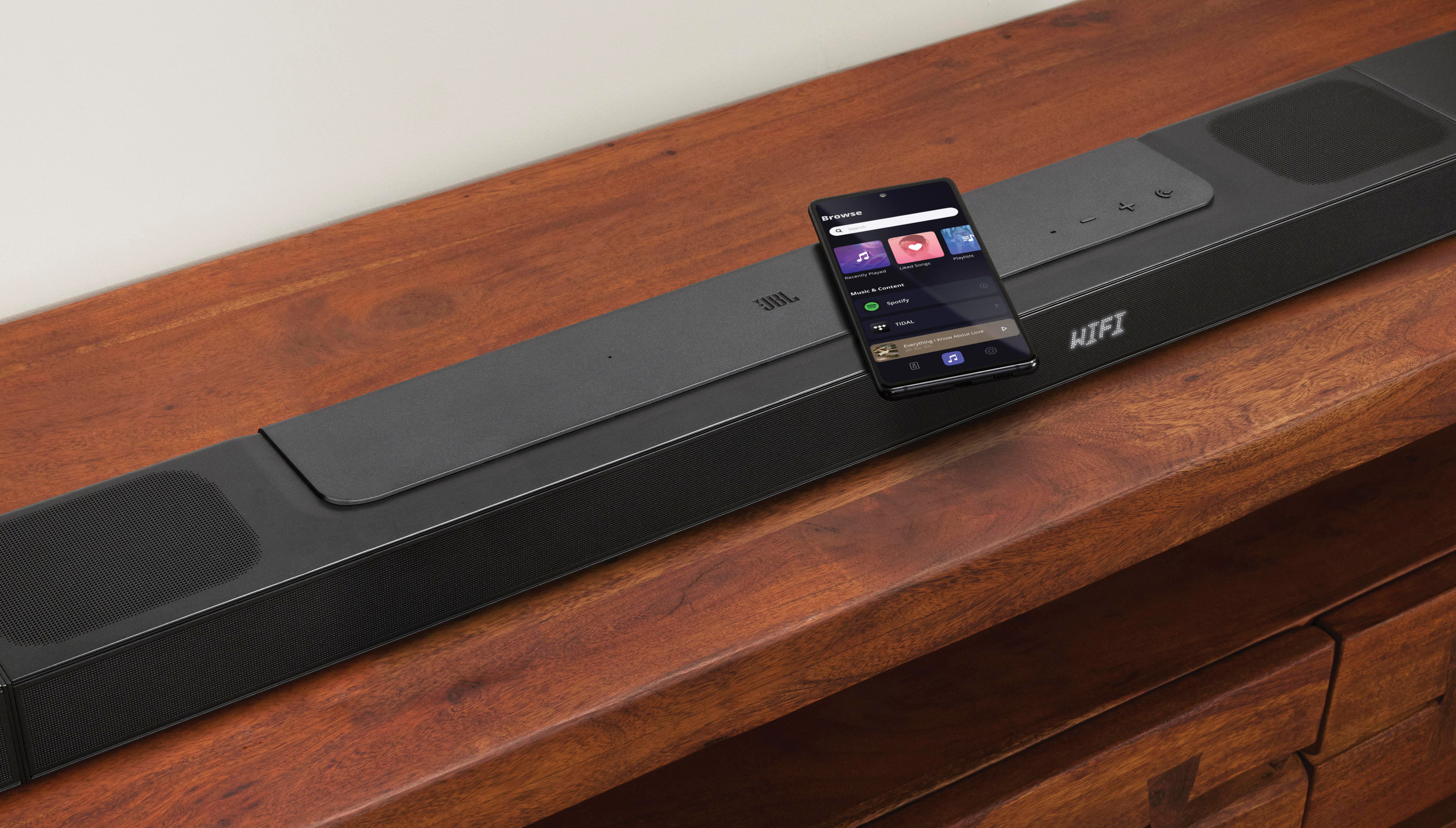Eine schwarze Soundbar mit einem Smartphone darauf. Das Telefon zeigt Musik-Apps an. Es liegt auf einer Holzoberfläche vor einer weißen Wand.