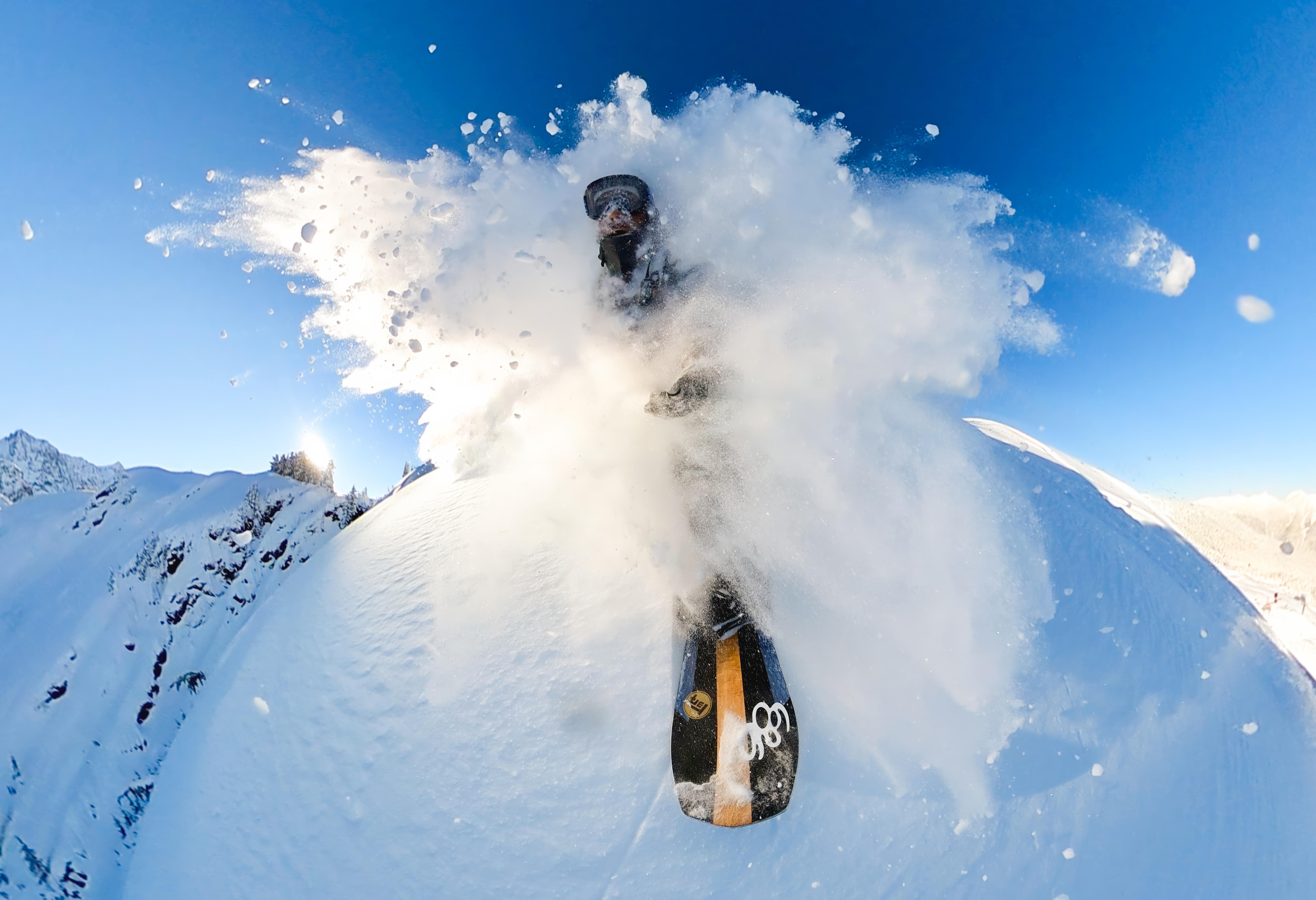 Ein Snowboarder fährt durch tiefen Pulverschnee und erzeugt eine große Wolke. Blauer Himmel im Hintergrund. Das Board ist sichtbar.