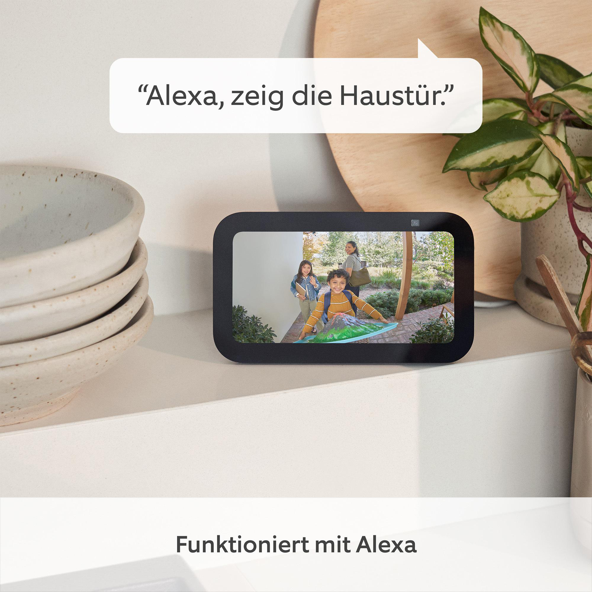 Ein Echo Show zeigt eine Familie an einer Tür mit Text: Alexa, zeig die Haustür. Pflanzen und Schalen stehen auf der Theke.