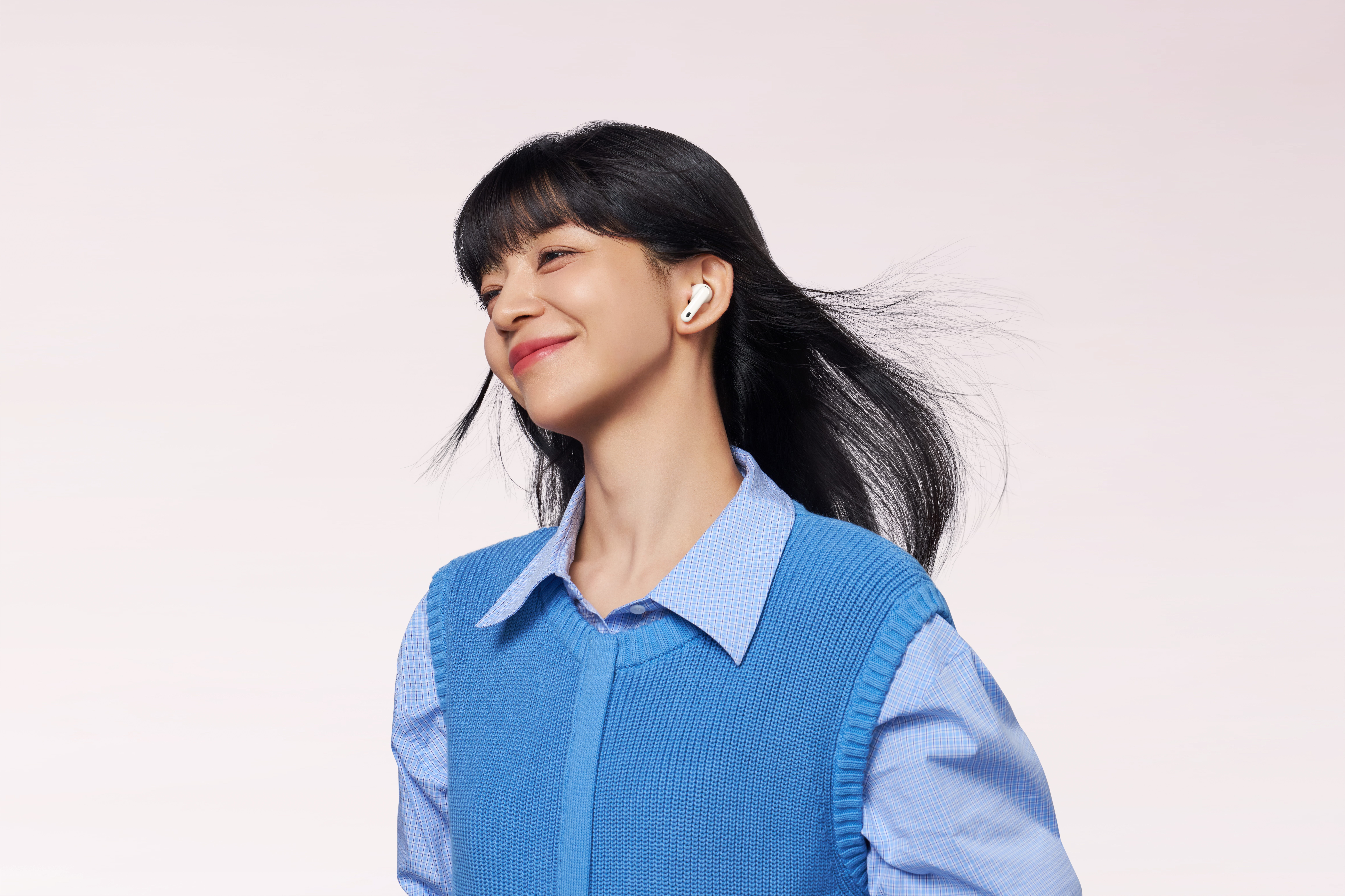 Uśmiechnięta kobieta z czarnymi włosami, niebieskim swetrem i koszulą, jedną słuchawką. Tło jest jasnoróżowe.