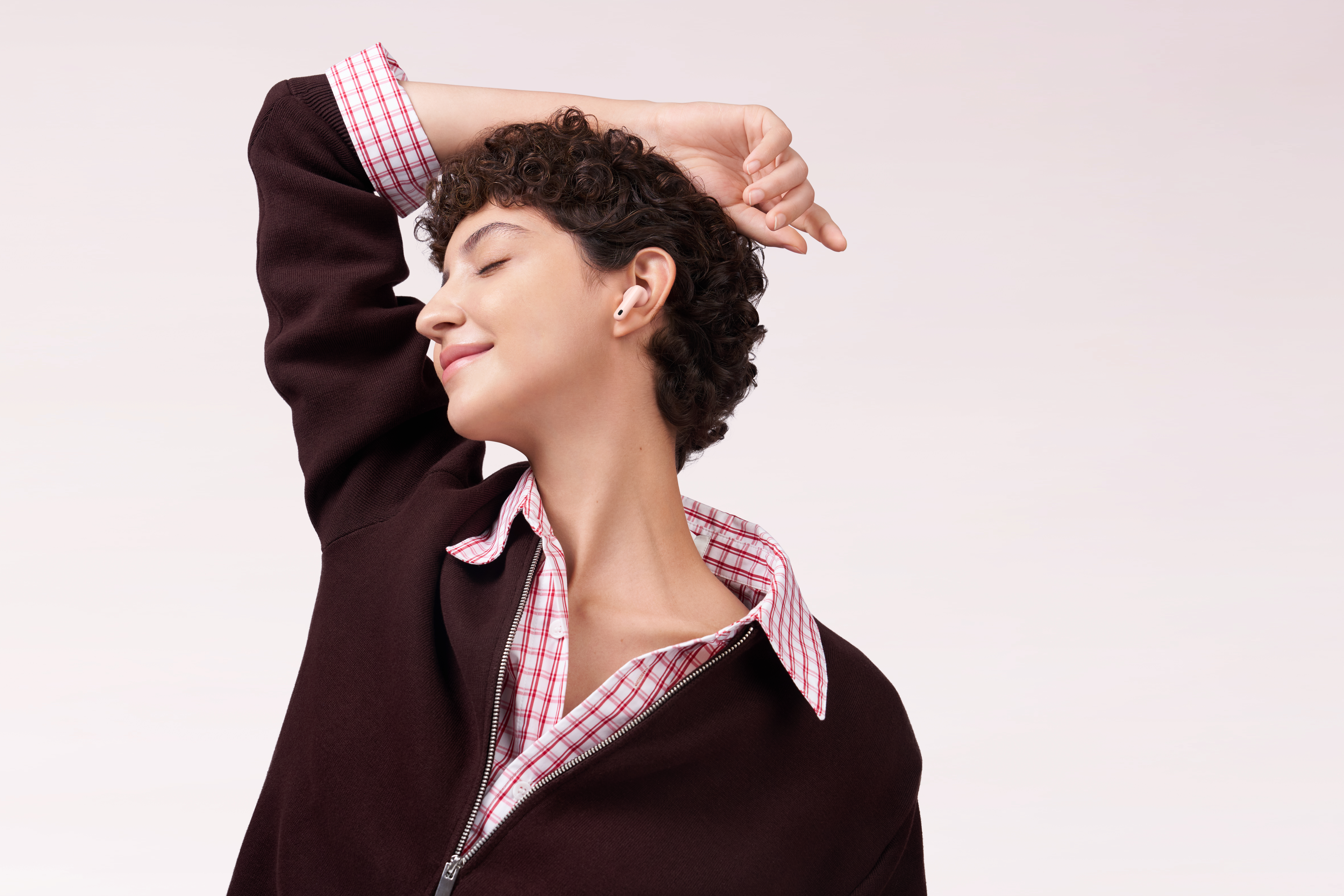 Kobieta z kręconymi włosami, w bordowej kurtce i koszuli w kratę, ze słuchawkami w uchu i zamkniętymi oczami.