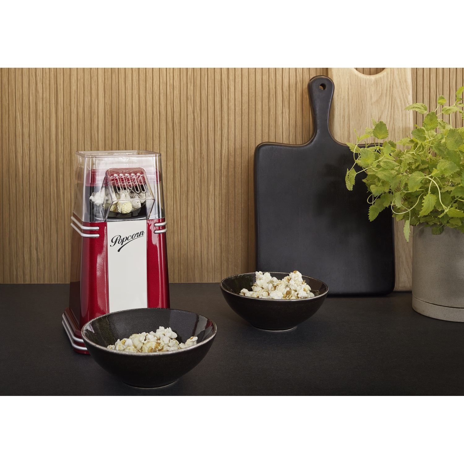 Una macchina per popcorn rossa con due ciotole di popcorn. Un tagliere nero, una pianta in vaso e sfondo a pannelli di legno.