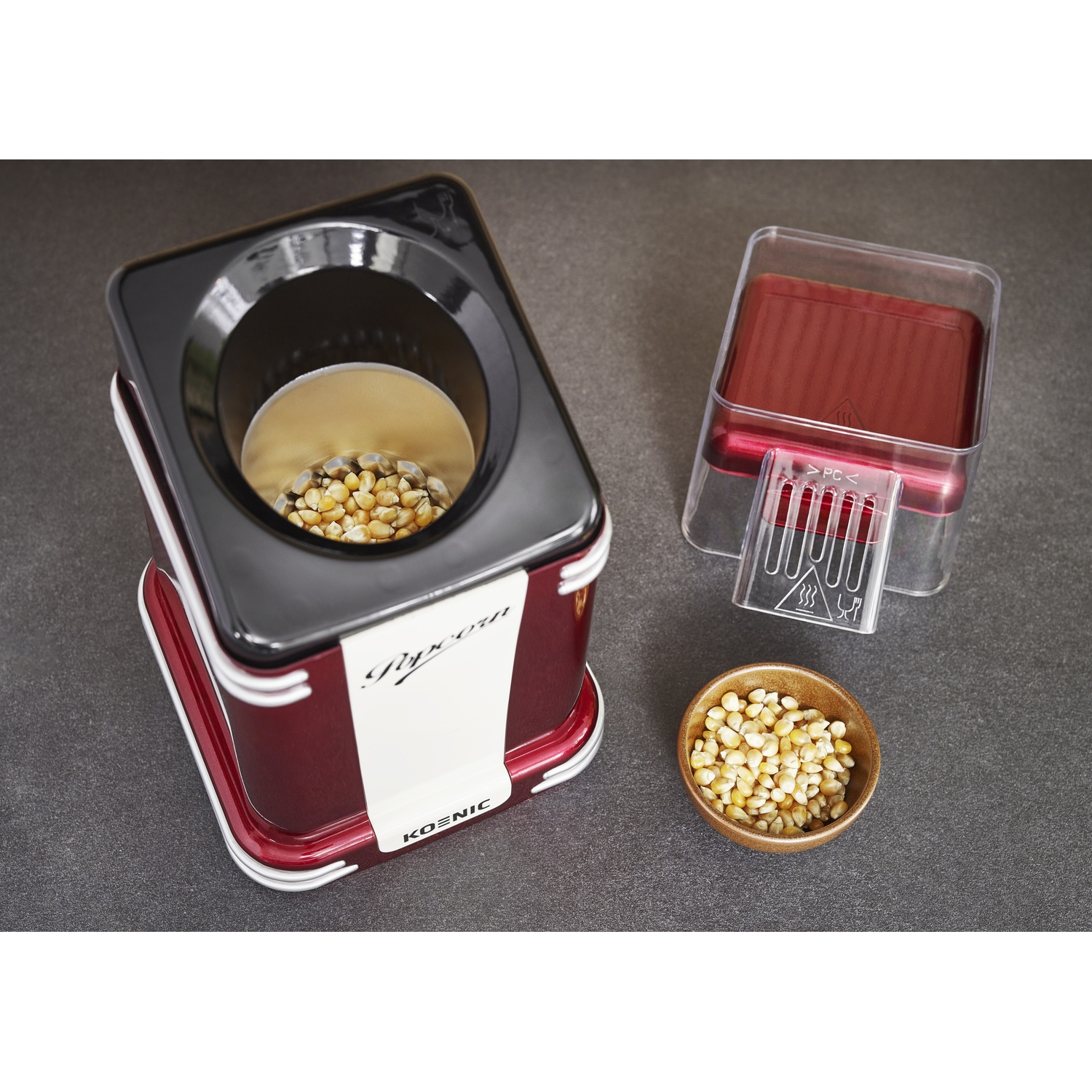 Una macchina per popcorn rossa e nera con chicchi di mais all'interno e una piccola ciotola di chicchi. Il coperchio e il misurino sono vicini.