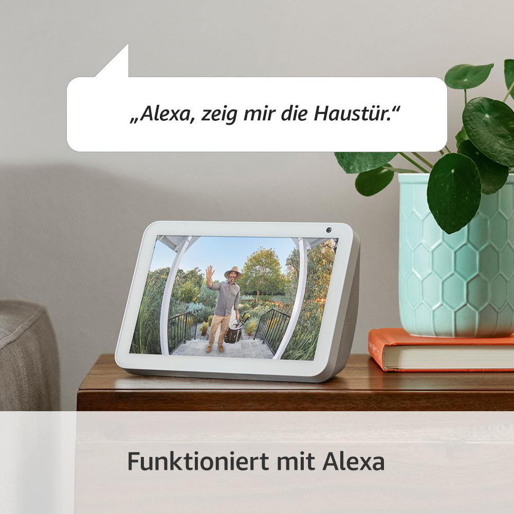 Ein Smart Display zeigt einen winkenden Mann. Textblase sagt 'Alexa, zeig mir die Haustür'. Pflanze und Buch rechts.