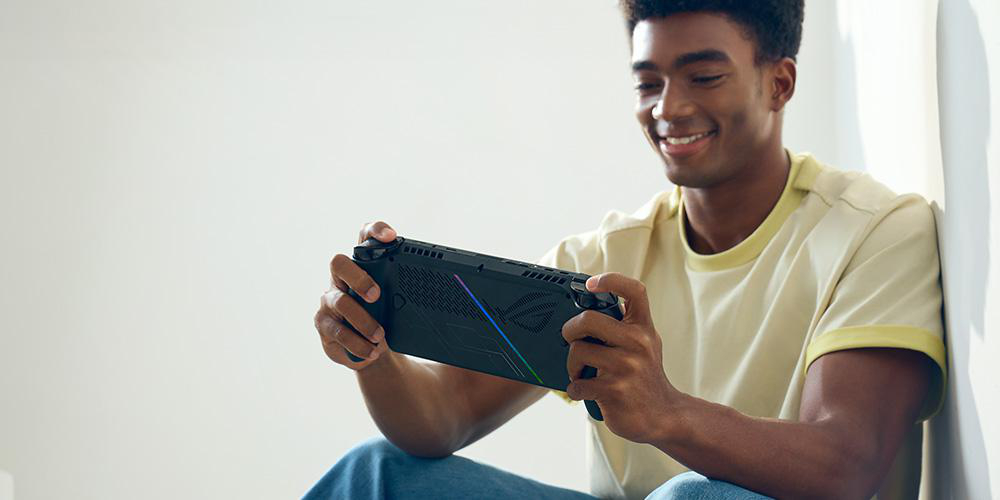 Ein Mann mit dunkler Haut lächelt, während er eine schwarze Handheld-Spielkonsole hält. Er sitzt auf hellem Hintergrund, trägt ein gelbes Hemd.