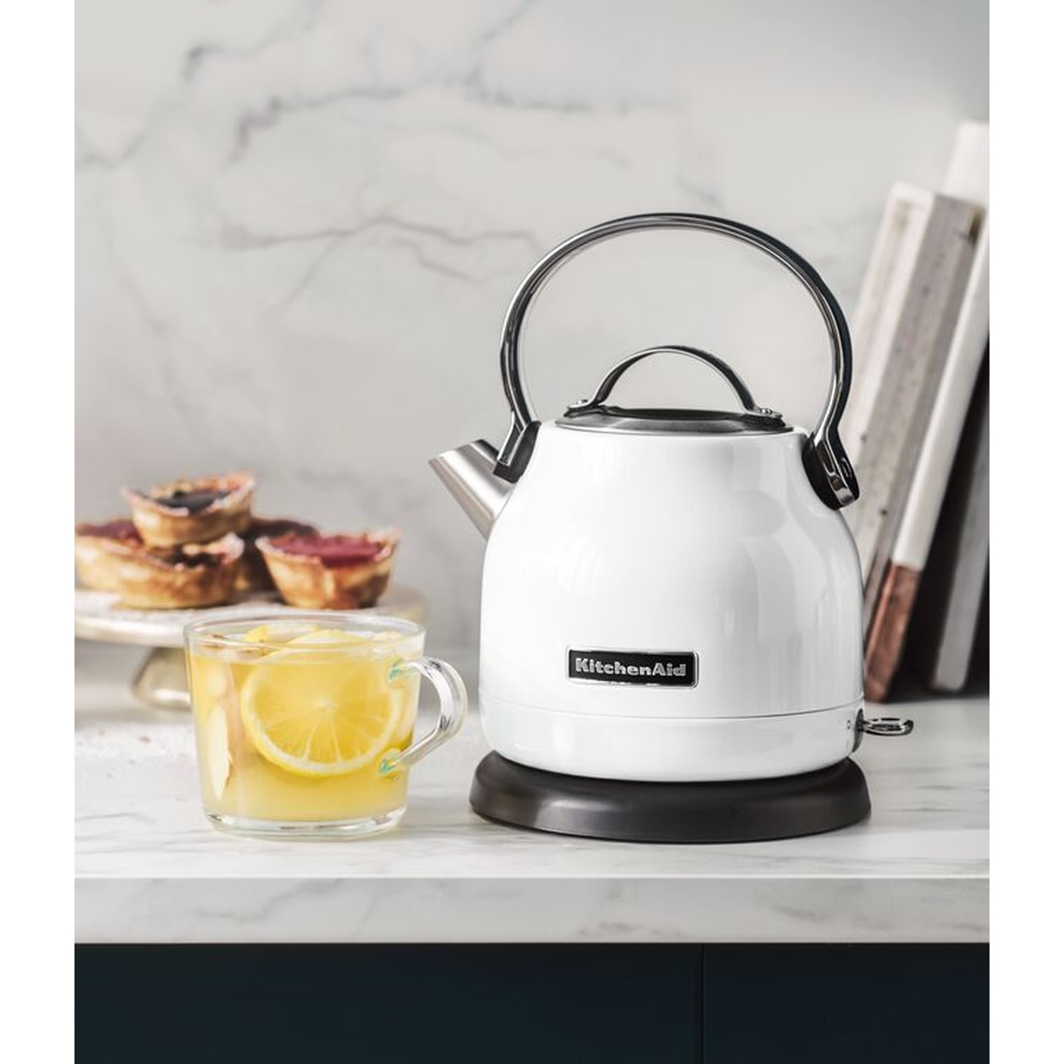 Un bollitore KitchenAid bianco accanto a un bicchiere di tè al limone. Un piatto di crostatine sullo sfondo. Piano di lavoro in marmo.