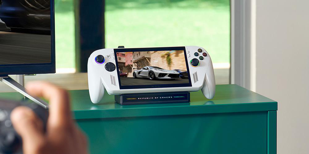 Eine weiße Handheld-Spielkonsole steht auf einem grünen Tisch und zeigt ein Autorennen. Eine Hand hält einen Controller.