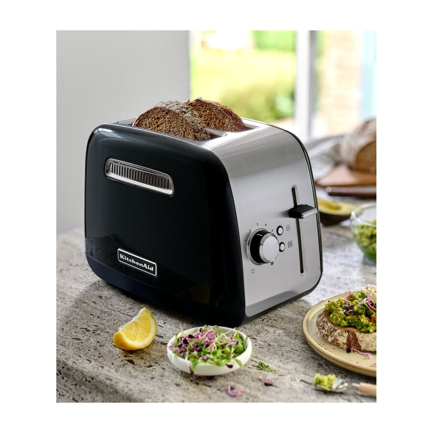 Un tostapane KitchenAid nero e argento con toast all'interno. Un piatto di toast con avocado, spicchio di limone e una ciotolina con verdure.