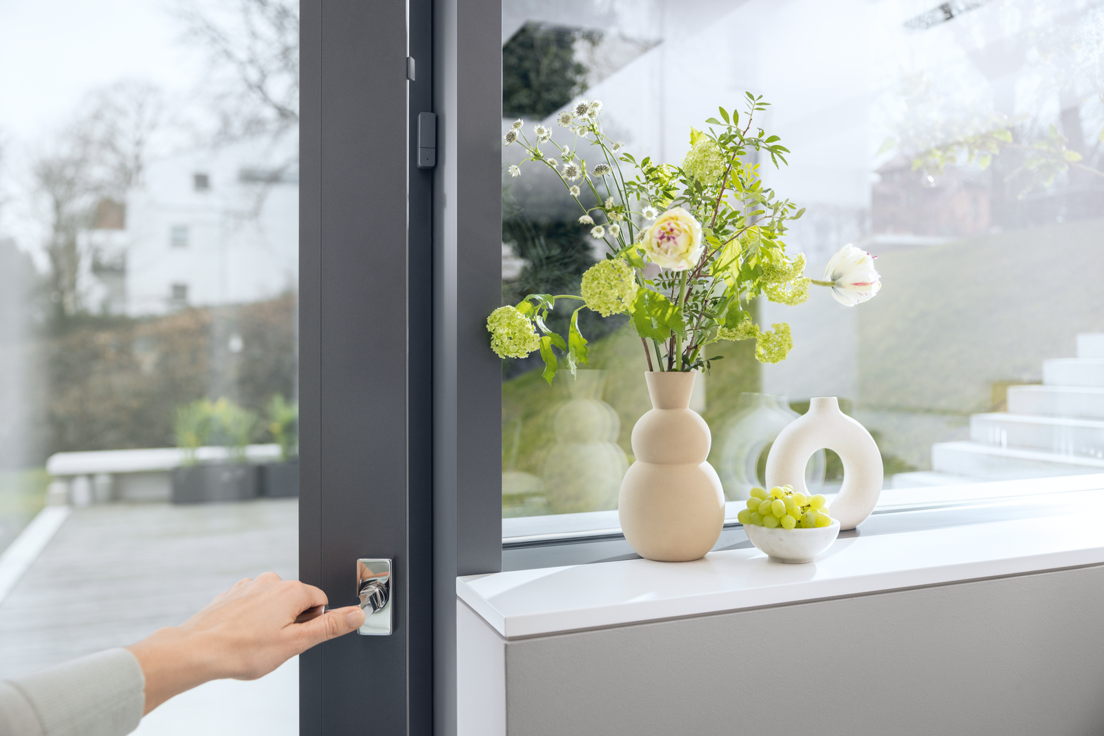 Eine Hand öffnet ein Fenster. Blumen in einer Vase stehen auf der Fensterbank, mit Trauben und einer dekorativen Vase. Draußen ist eine verschwommene Stadtlandschaft.