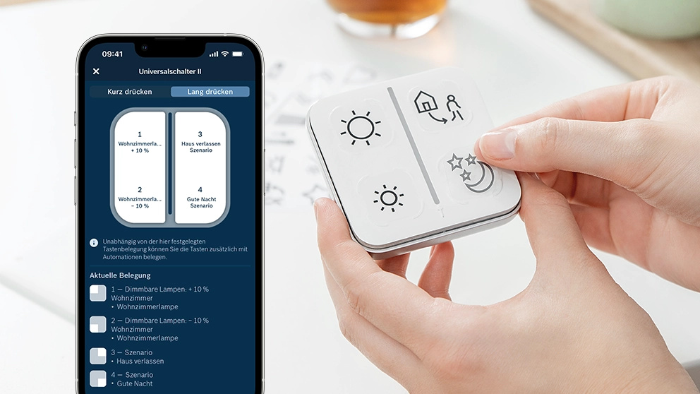 Eine Hand hält eine weiße Smart-Home-Fernbedienung. Ein Smartphone zeigt Einstellungen an. Die Fernbedienung hat Symbole für Sonne, Haus und Mond.