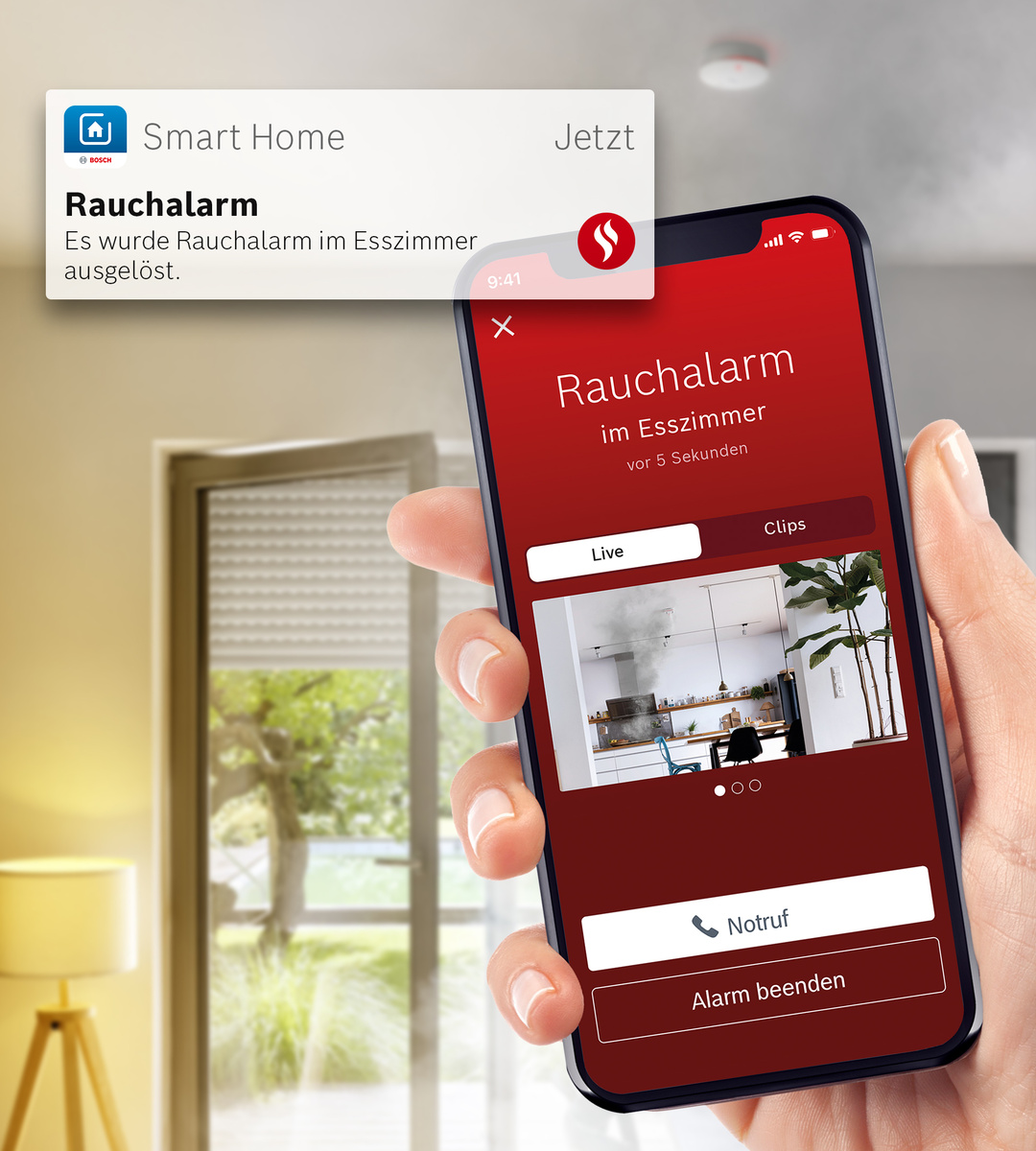 Eine Hand hält ein Telefon, das eine Rauchmelderbenachrichtigung anzeigt. Der Bildschirm zeigt eine Küche mit Rauch. Eine separate Benachrichtigung ist ebenfalls sichtbar.