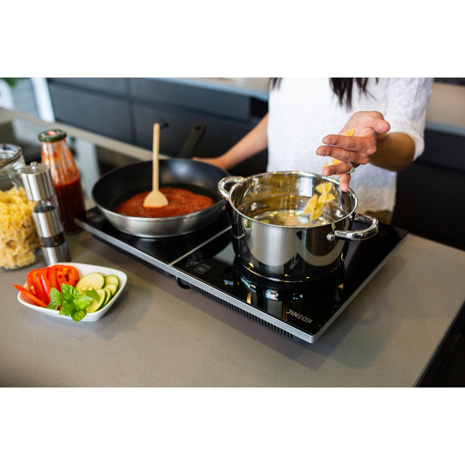 Una persona sta cucinando la pasta. Una padella con salsa e una pentola con acqua su un piano cottura nero. Verdure e ingredienti a lato.